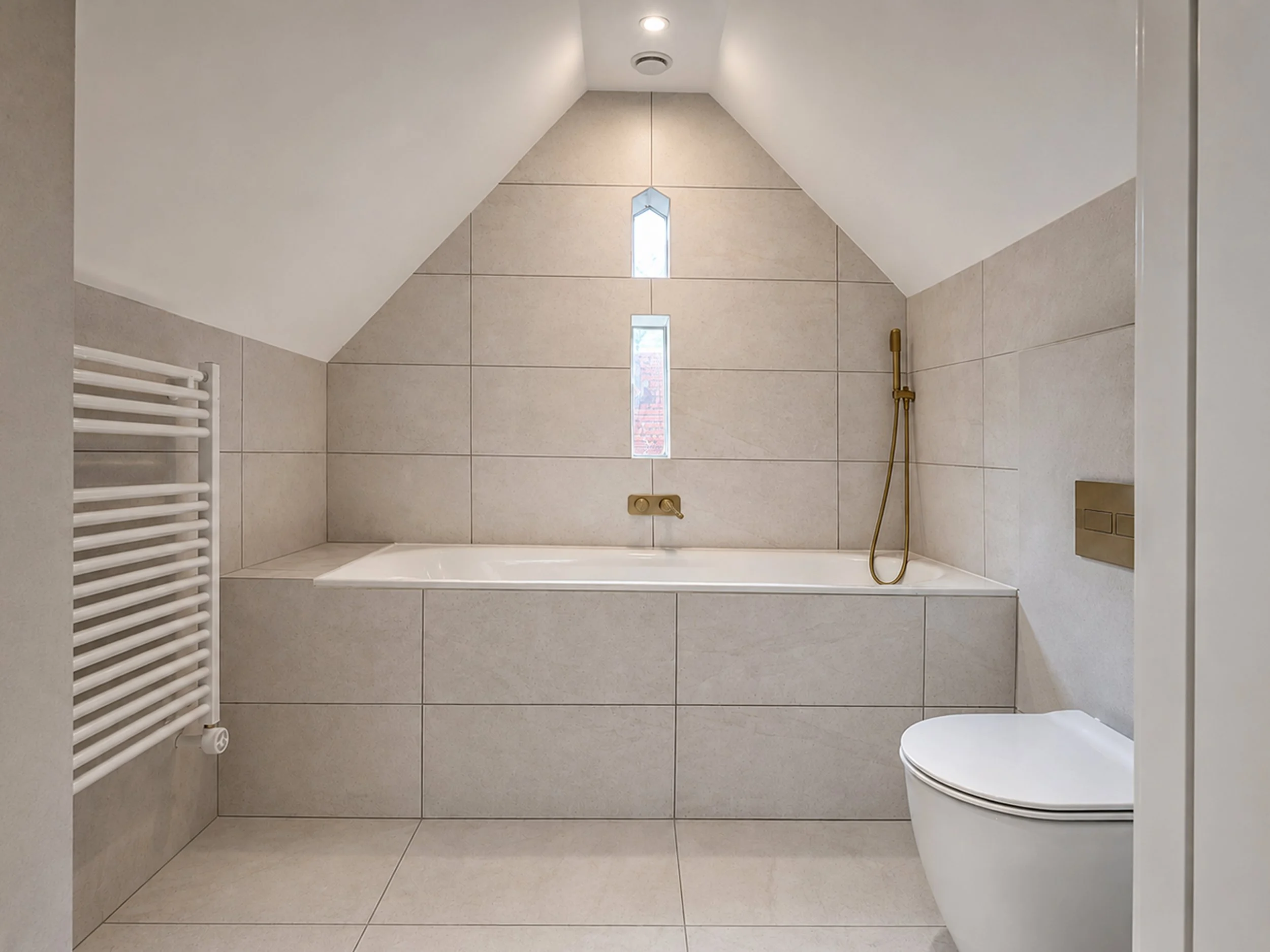 A beautiful minimalist bathroom in East Peckham with integrated storage, grey panelling, bath with brushed brass fixtures, shower and white toilet, bath and towel rail. loft conversion for a barn in Kent by Studio Jetset in Faversham