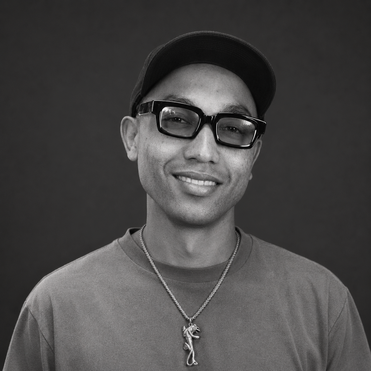 Black and white portrait of a smiling man wearing a cap, thick glasses, a T-shirt, and a necklace with a seahorse pendant.