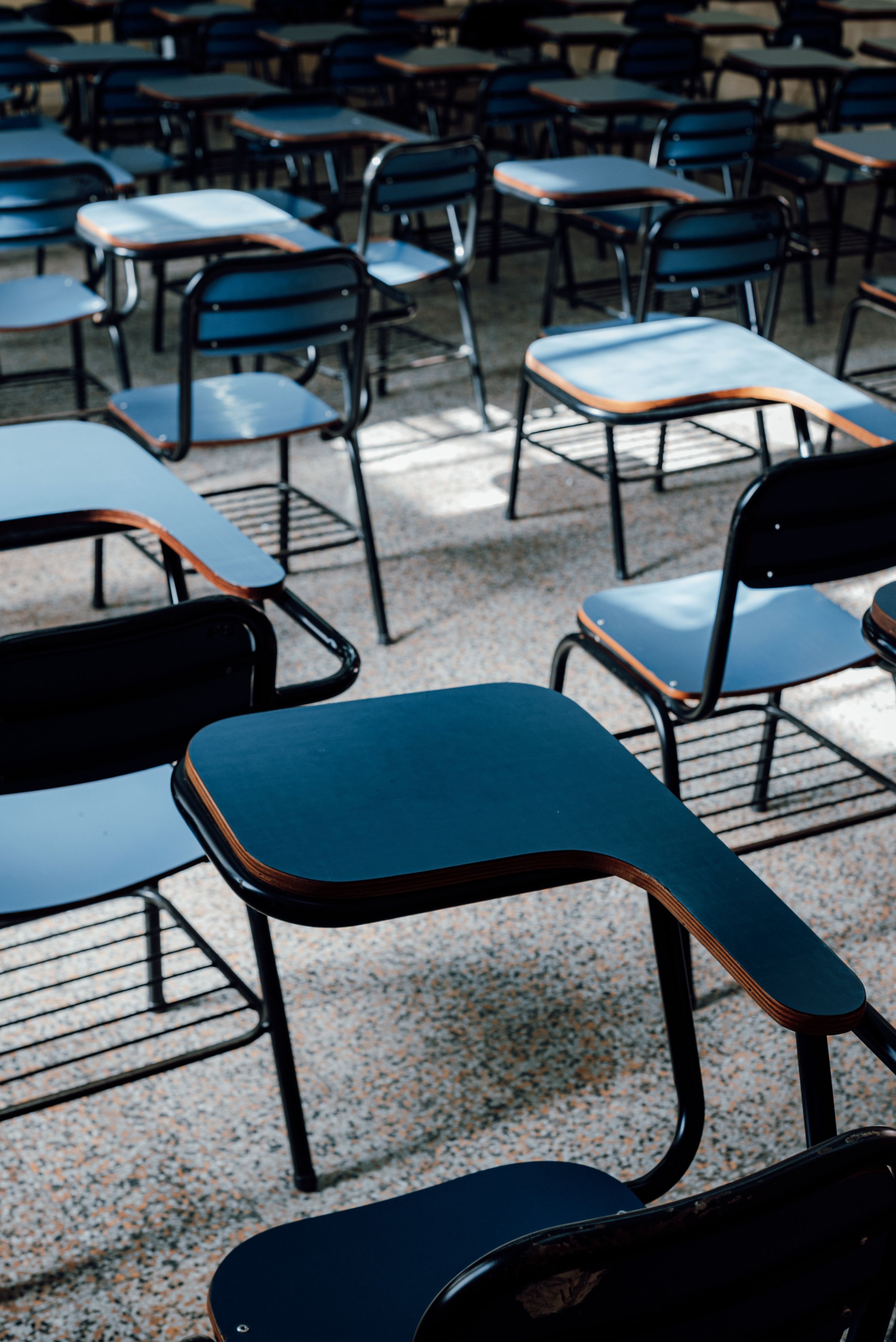Multiple empty school desks with black metal frames and blue tabletops arranged on a terrazzo floor, with sunlight casting shadows.