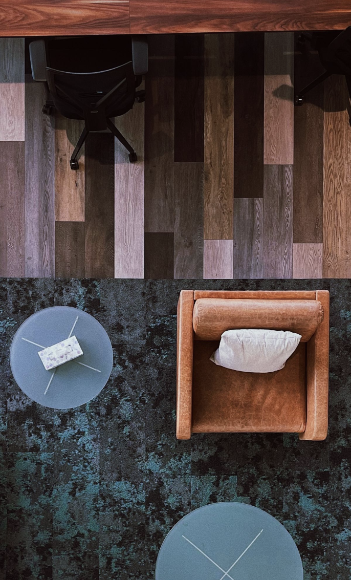 Overhead view of a workspace with a wooden desk, an ergonomic office chair, a small round glass table with a tissue box, and an orange armchair with a pillow, set on a patterned dark blue carpet.