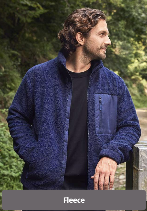 A man with wavy brown hair and a beard wearing a navy fleece jacket over a black shirt, standing outdoors with green trees in the background.
