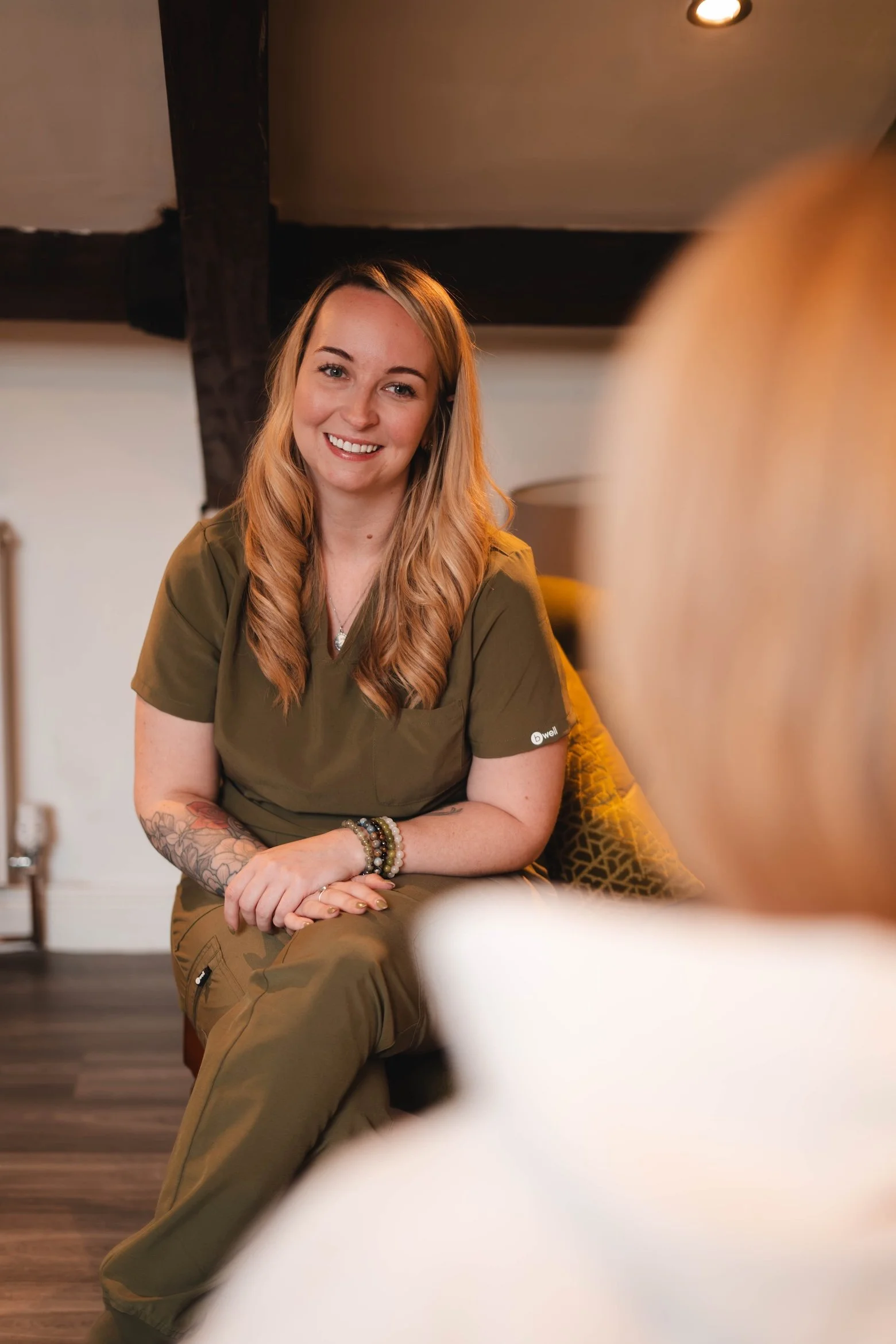 A smiling woman with long, wavy blonde hair and tattoos on her left arm, wearing green medical scrubs, sitting in a cozy room with dark wood beams on the ceiling and a soft-lit background, engaging in conversation with a blurred person in the foreground.