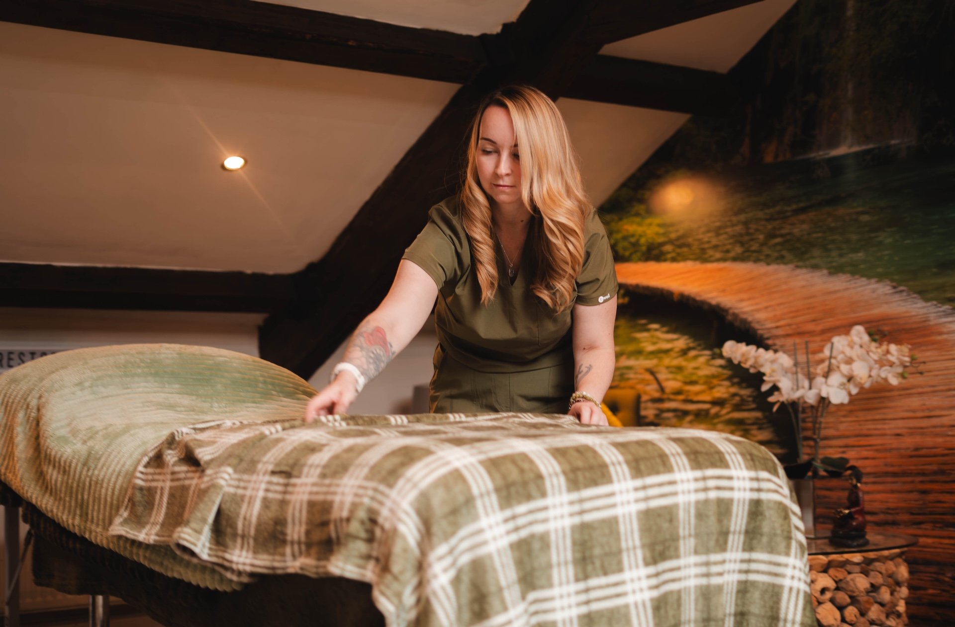 A woman with long blonde hair wearing a green uniform making a bed in a room with a nature-themed mural on the wall, a side table with a vase of white flowers, and wooden beams on the ceiling.