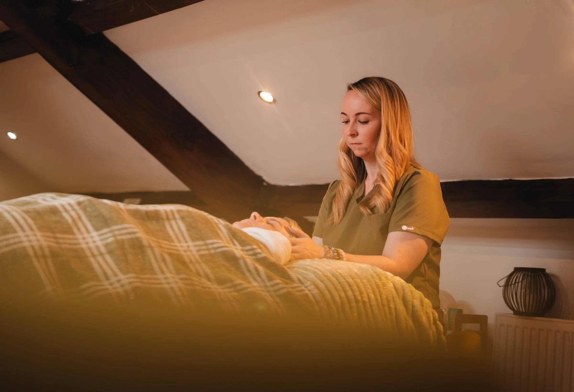 A woman with long blonde hair and a green scrub top stares at a baby lying on a bed covered with a beige and brown plaid blanket. The setting appears to be a cozy, warmly lit room with a sloped ceiling and wooden beams.