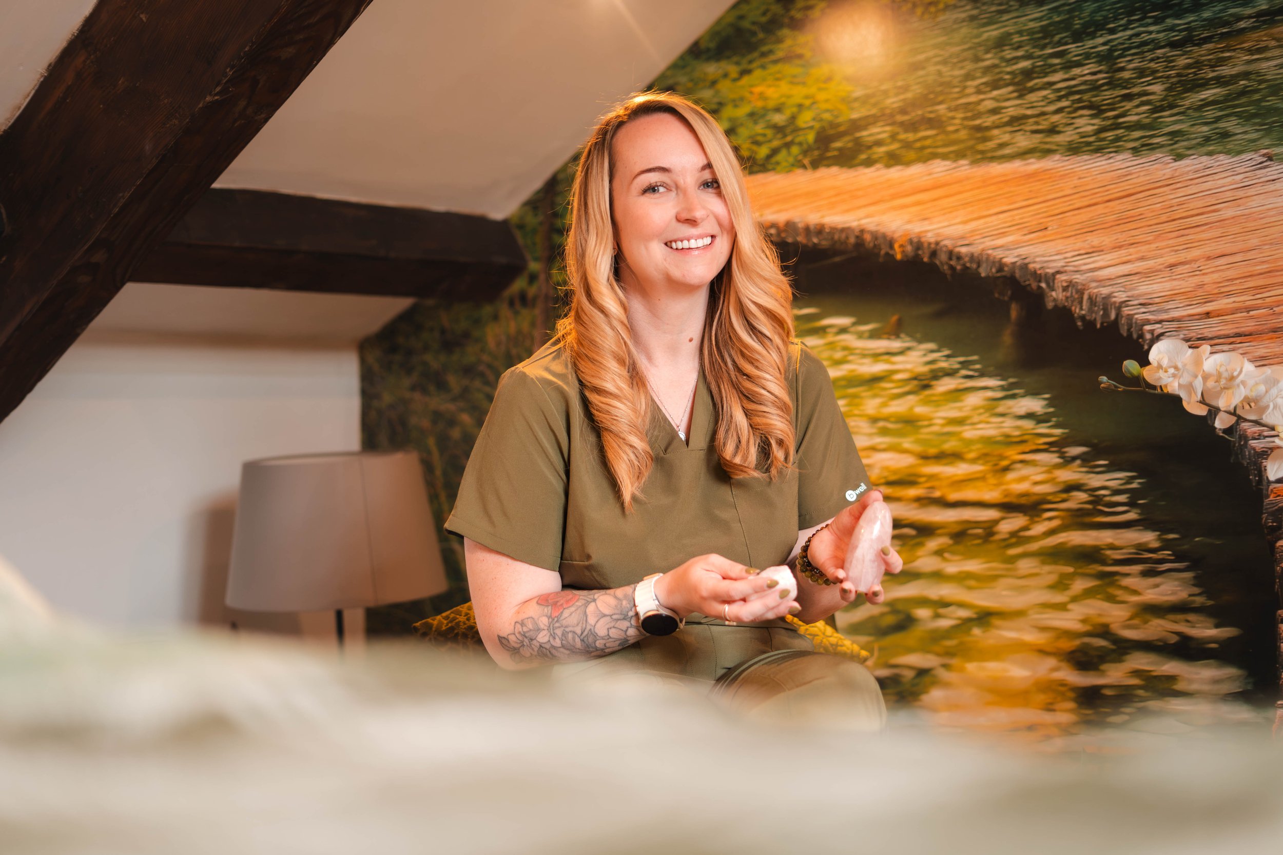 A woman with long, wavy blonde hair, tattoos on her arms, wearing an olive-green scrub top, smiling while holding crystals in her hands, sitting in front of a nature-themed mural featuring a wooden dock extending over a green lake.