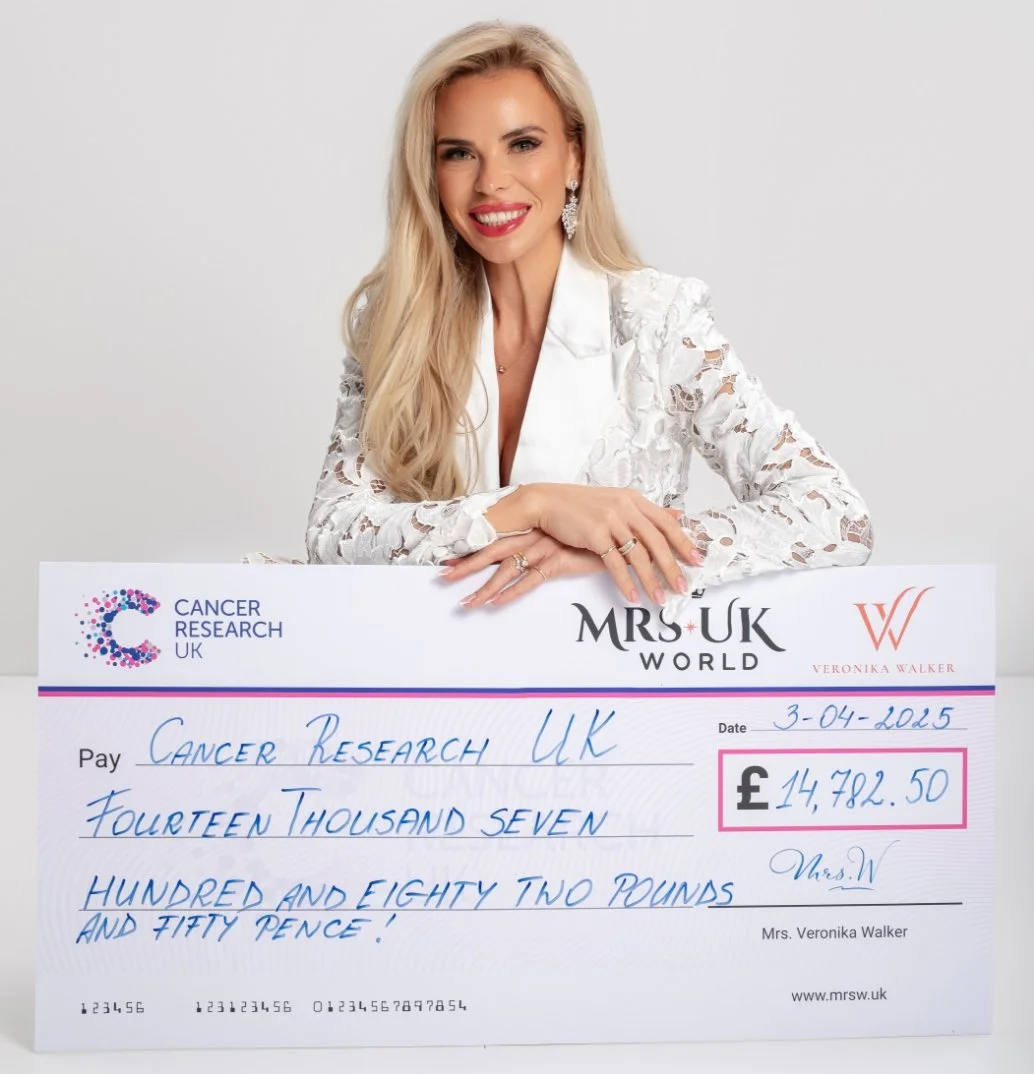 A woman with long blonde hair, wearing a white lace dress, smiling and sitting behind a large check. The check is made out to Cancer Research UK for 14,782.50 pounds, dated April 3, 2025, and signed by Mrs. Veronika Walker.