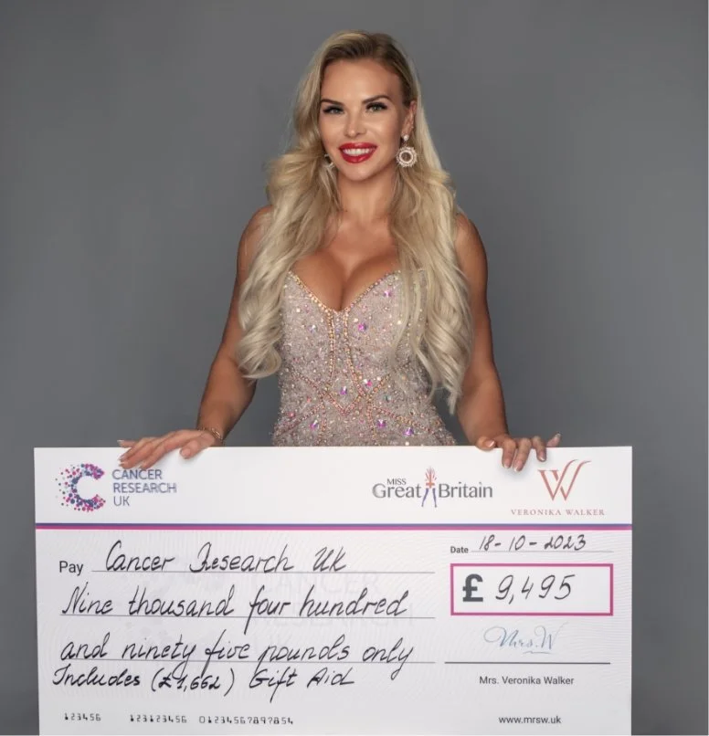 A woman with long blonde hair, wearing a sparkly dress and earrings, holding a large check for 9,495 pounds made out to Cancer Research UK, with logos of Miss Great Britain and Mrs. V.