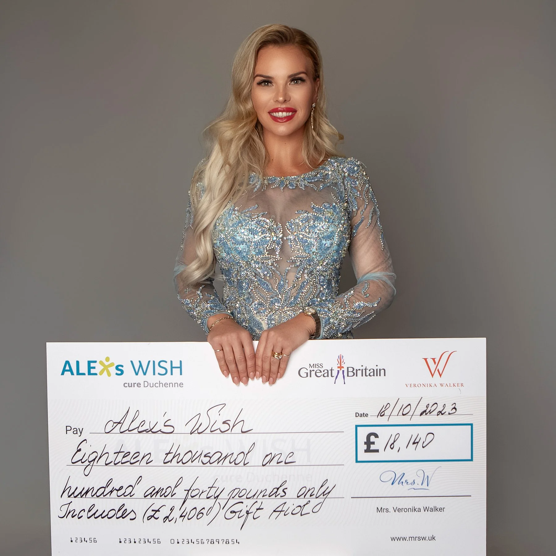 A woman in a silver embellished dress holds a large check made out to Alex's Wish for £18,140, dated 16/10/2023, issued by Mrs. Veronika Walker.
