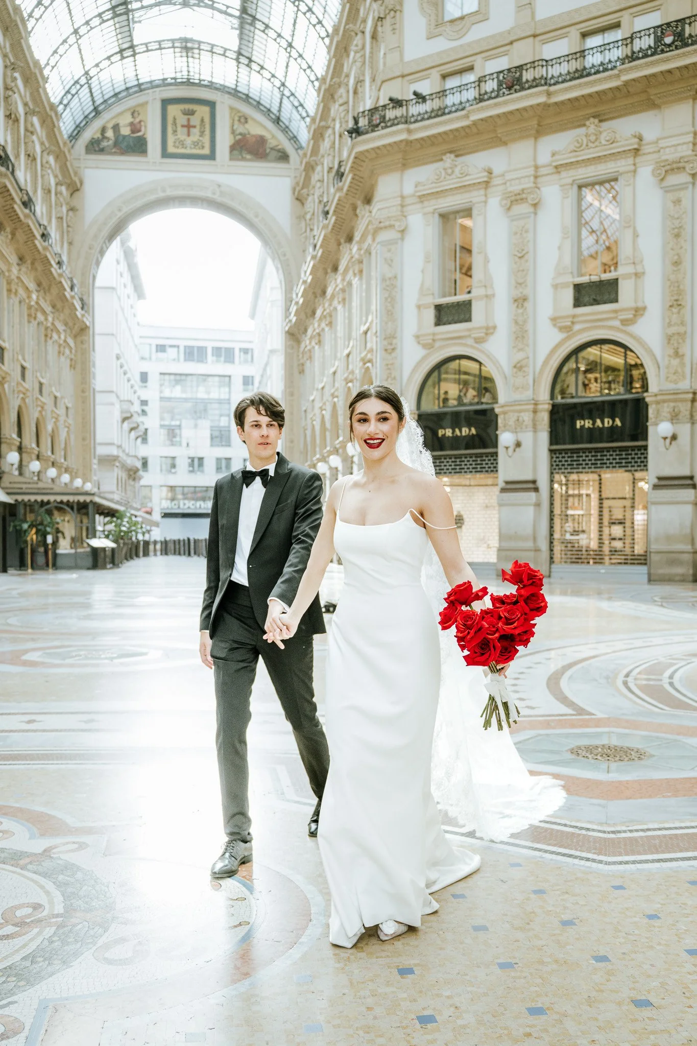 A bride and groom walking hand in hand inside a grand shopping arcade, with the bride holding a bouquet of red roses.