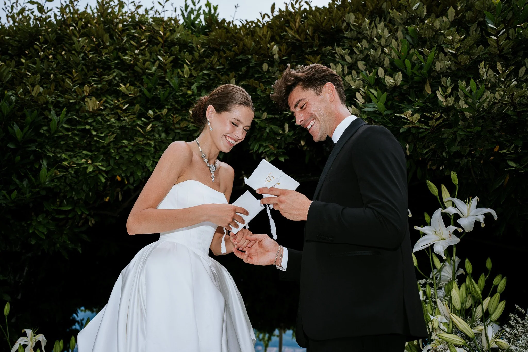 A bride and groom exchanging wedding vows outdoors, with the bride wearing a white strapless dress and diamond jewelry, and the groom in a black tuxedo, against a background of green hedge and white flowers.
