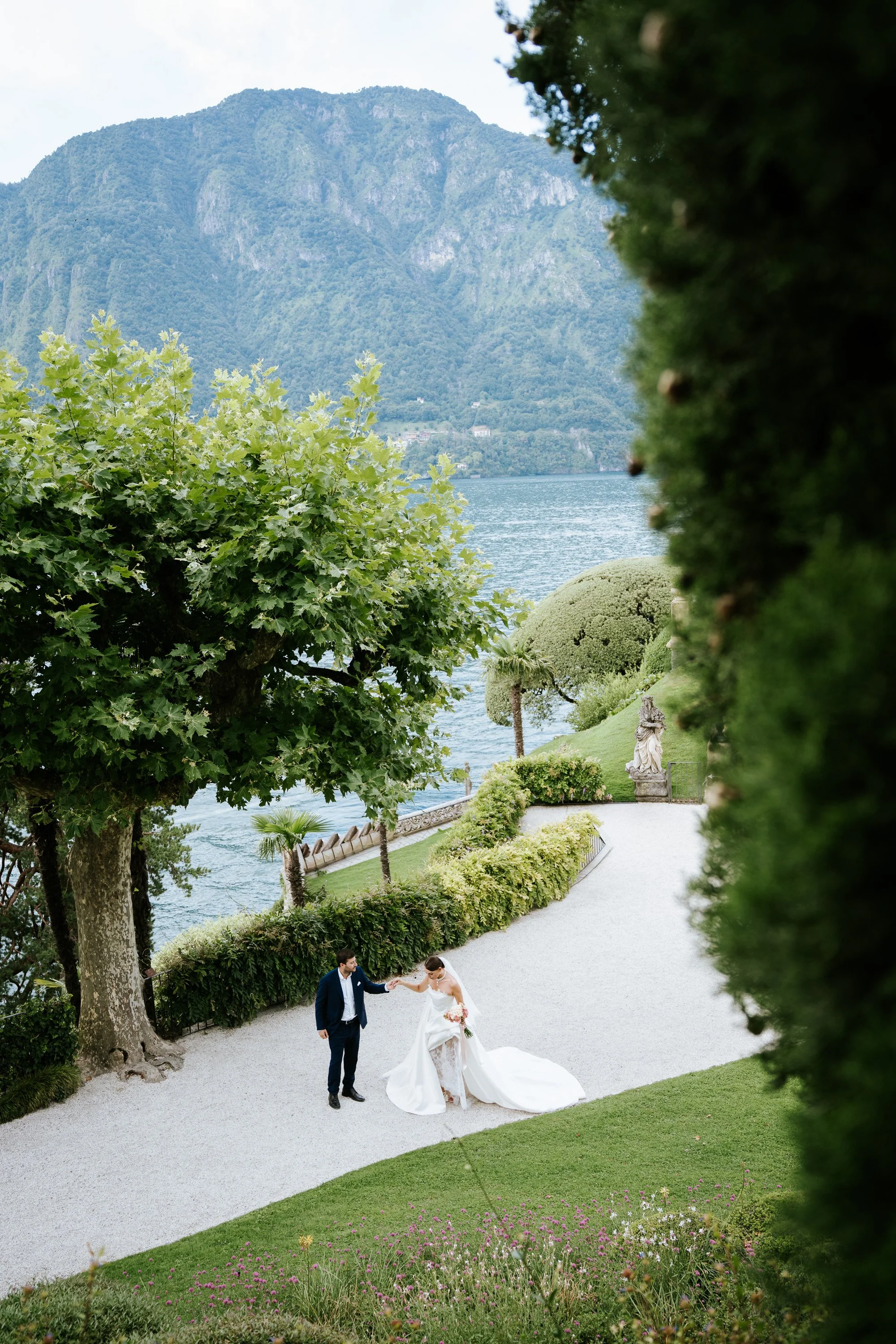 Bride and groom walking the garden path at Villa del Balbianello, Lake Como, past a stone statue.
