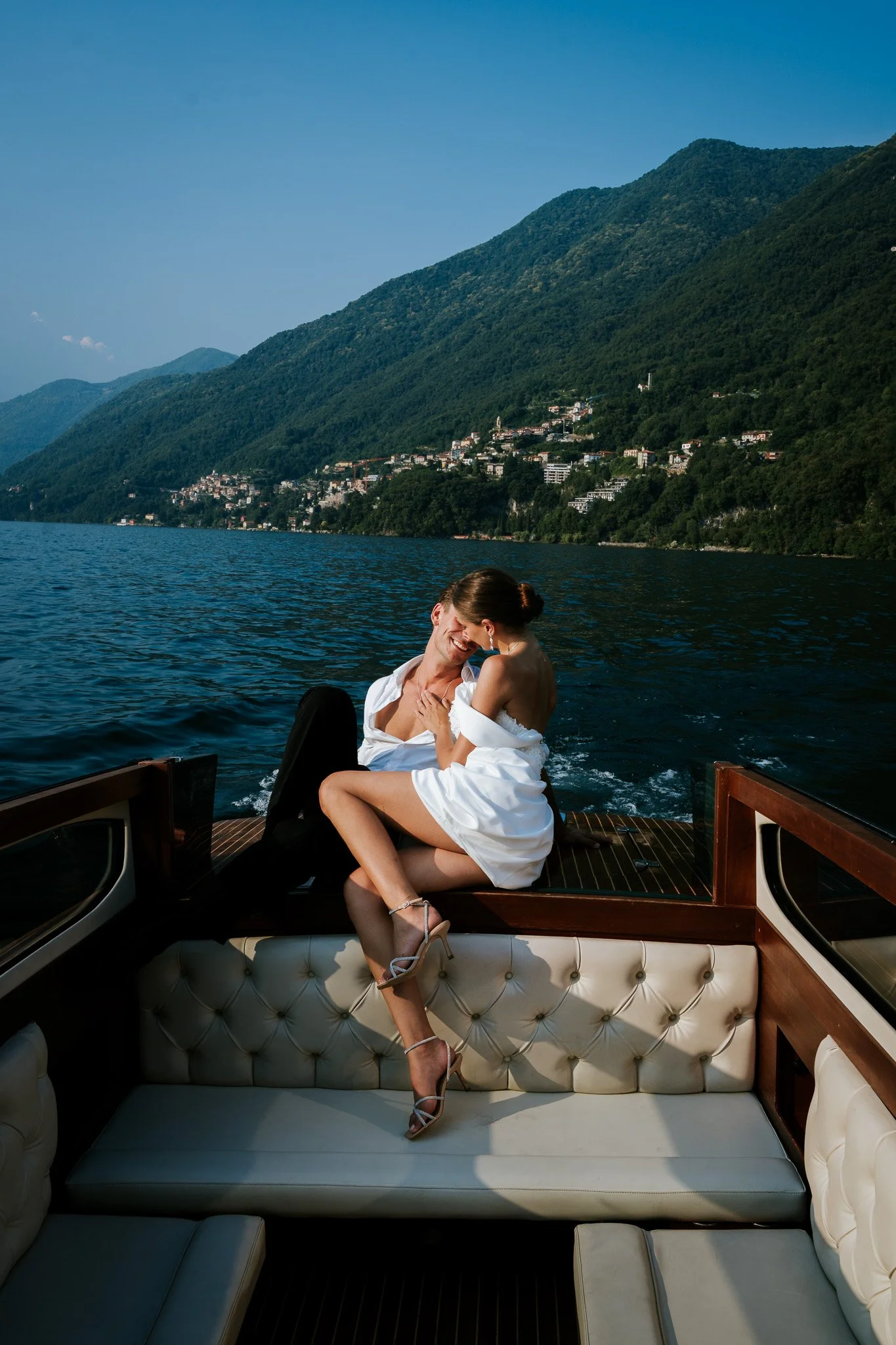 A couple embracing and smiling on the back of a boat during a sunny day, with a scenic view of green mountains and a lakeside town in the background.
