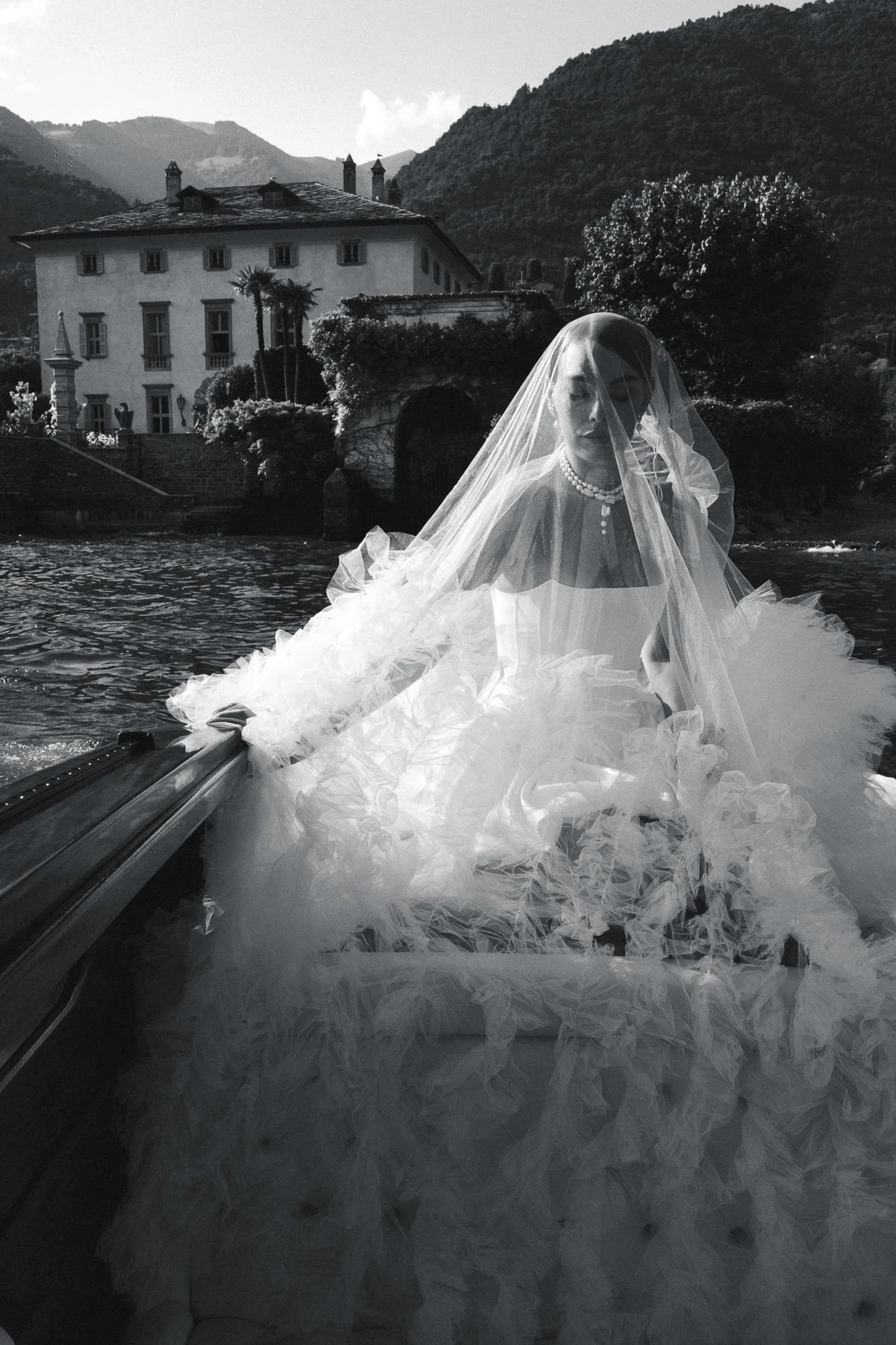 Black and white photo of a bride in her veil on a boat on Lake Como, with Villa del Balbiano and the mountains behind her.