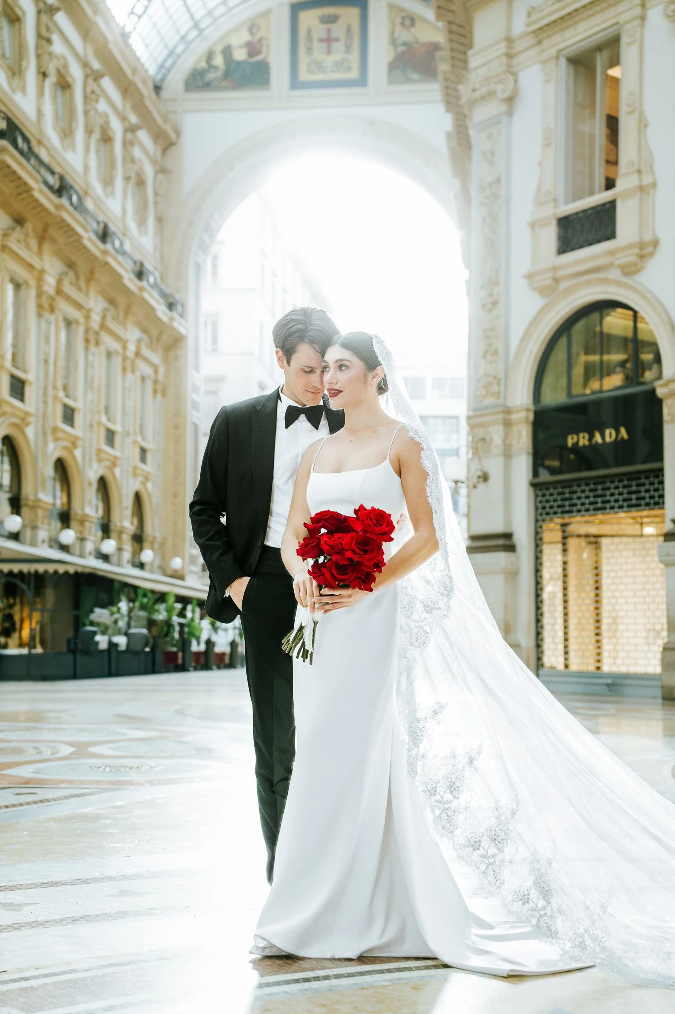 A bride and groom in wedding attire stand close together inside a grand shopping arcade with ornate architecture. The bride holds a bouquet of red roses, and the groom wears a tuxedo with a bow tie.