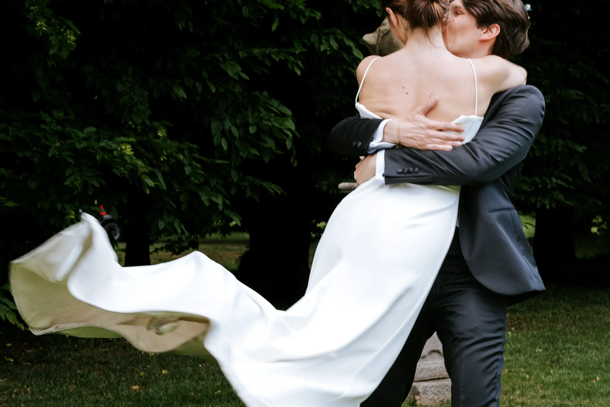 A couple dancing outdoors surrounded by green trees, with the woman wearing a white dress and the man in a black suit.