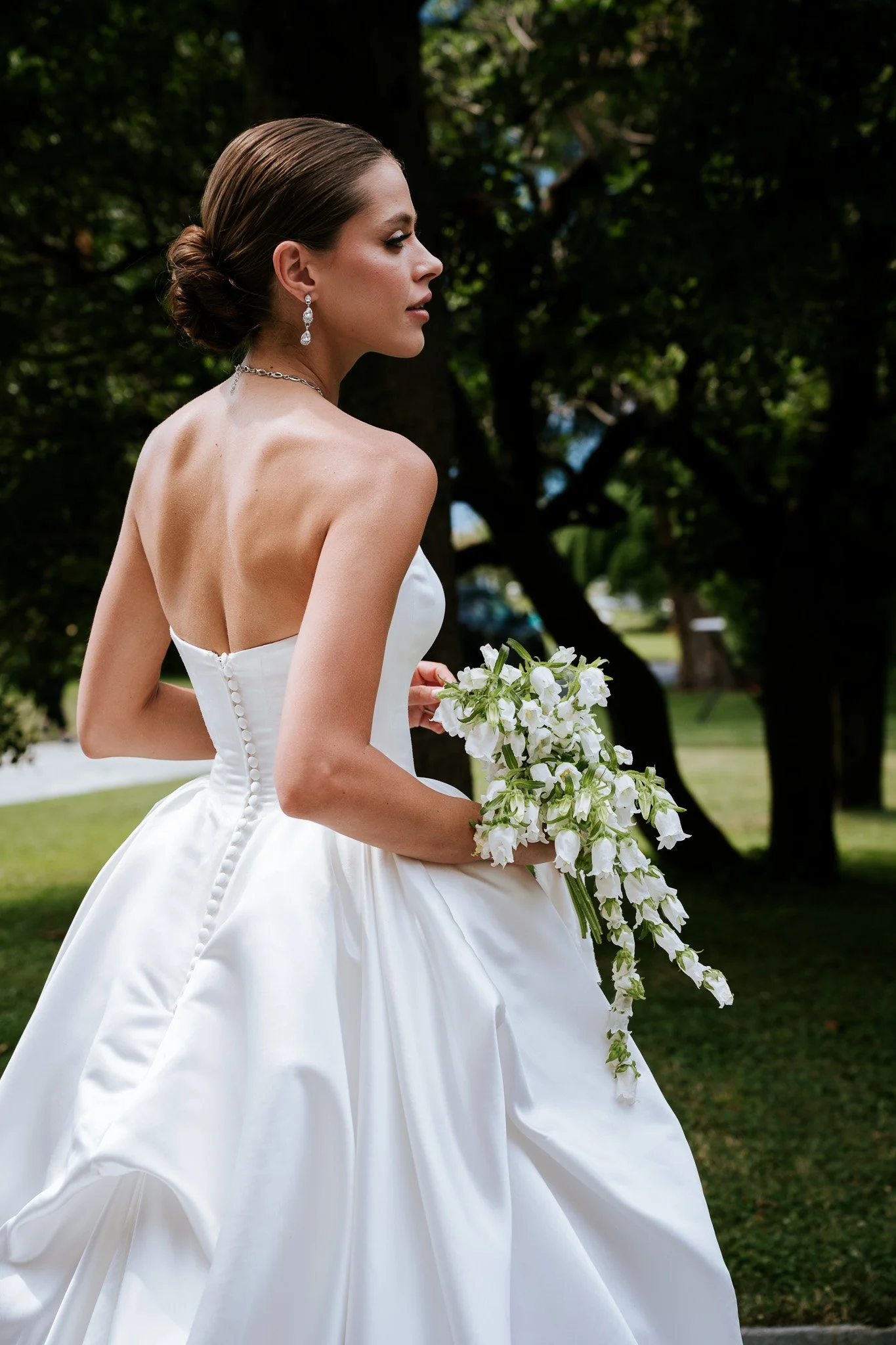 A bride in a white wedding gown holding a bouquet of white flowers outdoors with green trees in the background.