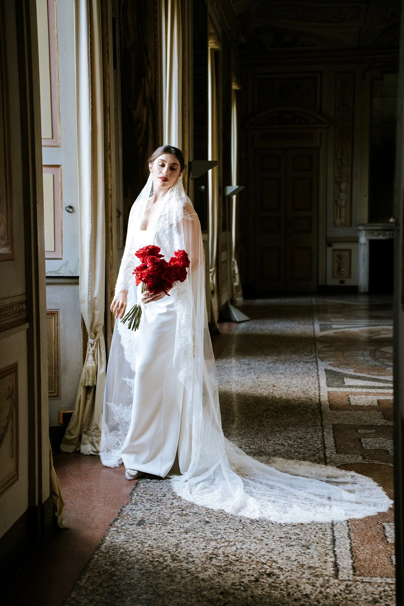 A bride in a wedding dress holding a bouquet of red roses, standing in a decorated, elegant interior with tall curtains and patterned carpet.