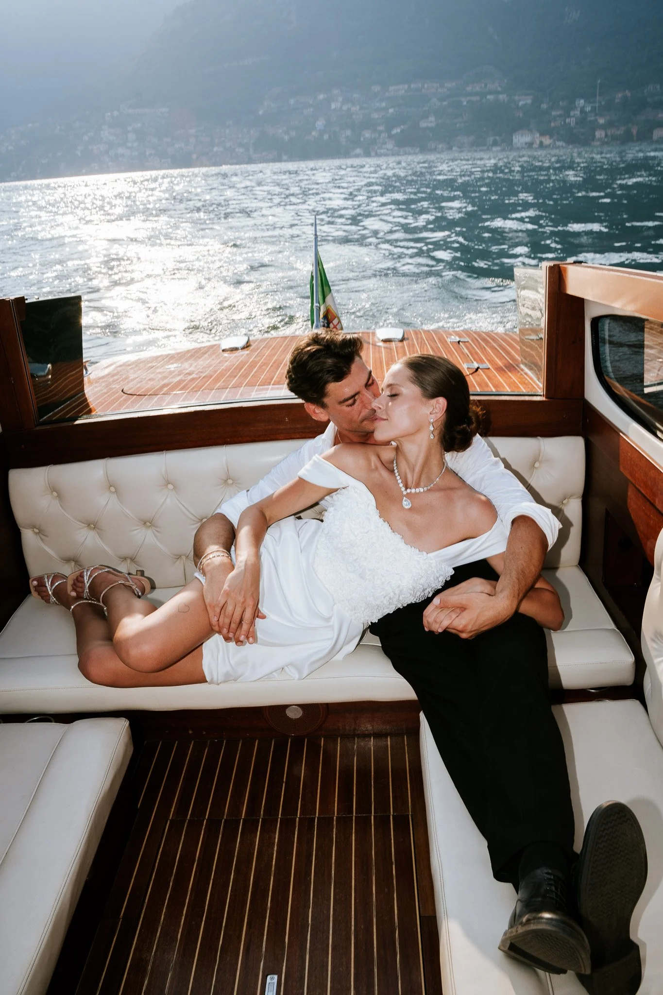 A man and a woman embrace and kiss on a yacht, with water and hills in the background. The woman is in a white dress and jewelry; the man is in black pants and a white shirt. They are sitting on a white cushioned bench inside the yacht.