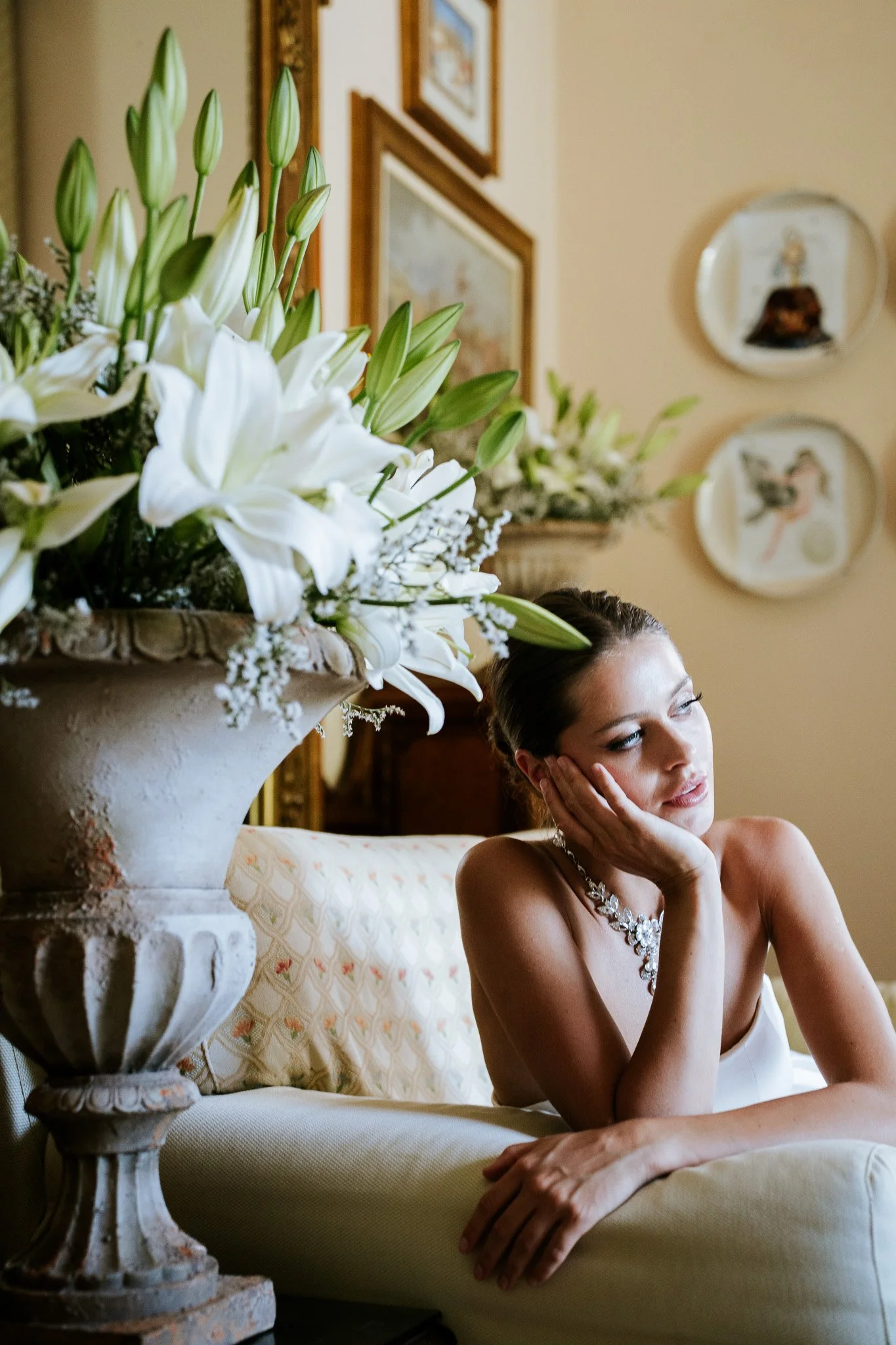 A woman sitting on a couch with her hand on her face, wearing a white dress and a floral necklace, next to a large vase with white lilies and buds, with framed artwork on the wall behind her.