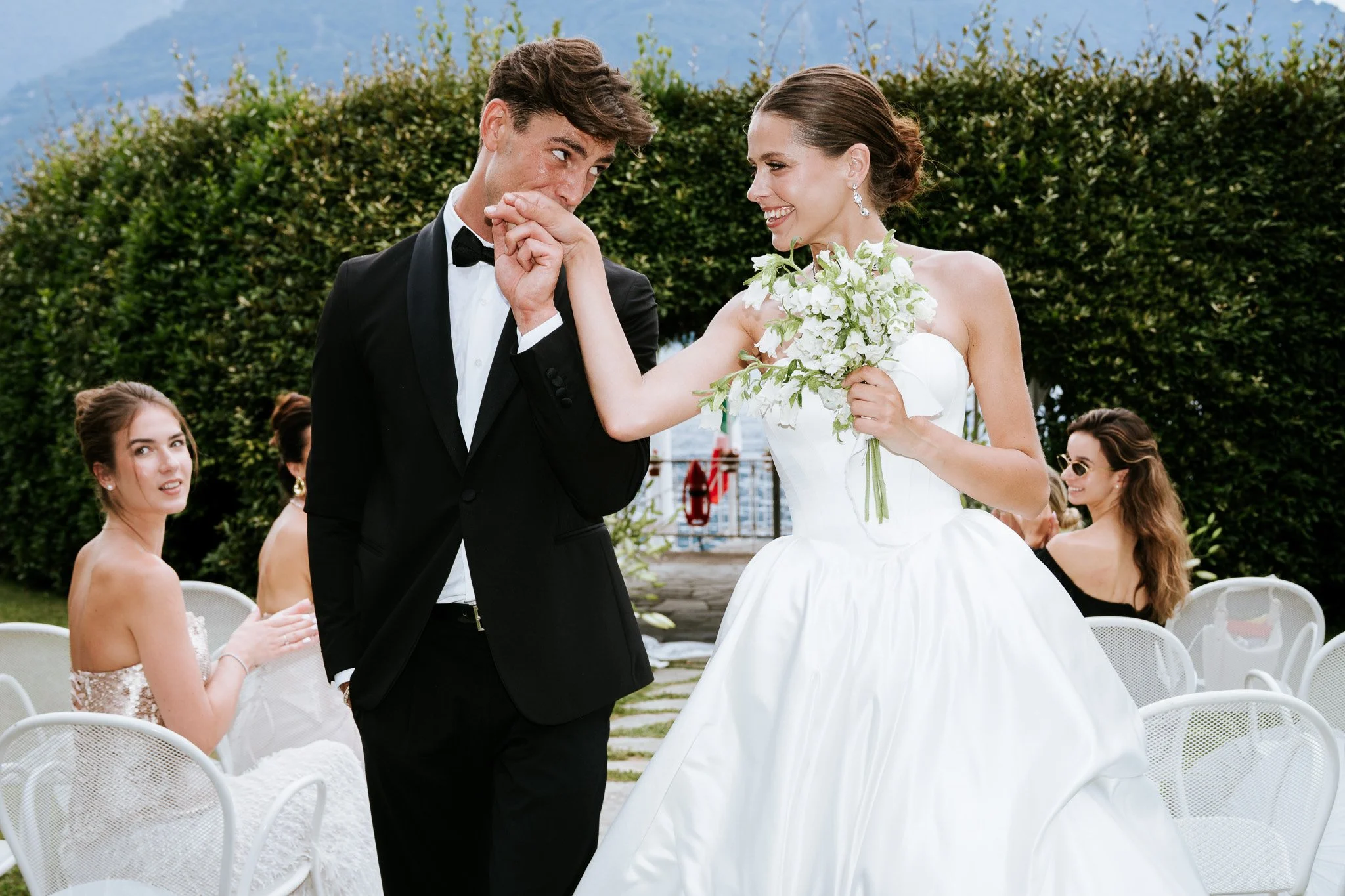 A bride and groom are dancing at their outdoor wedding, surrounded by seated guests, with a lush green hedge and mountains in the background.