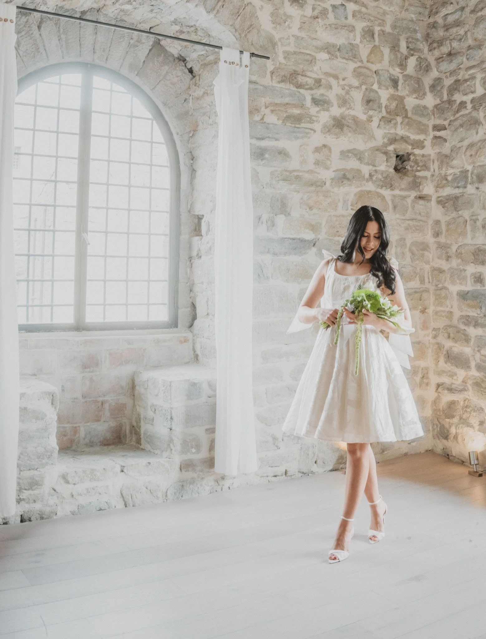 A young woman in a white dress standing in front of a stone wall with a large arched window. She is holding a bouquet and wearing high heel shoes.