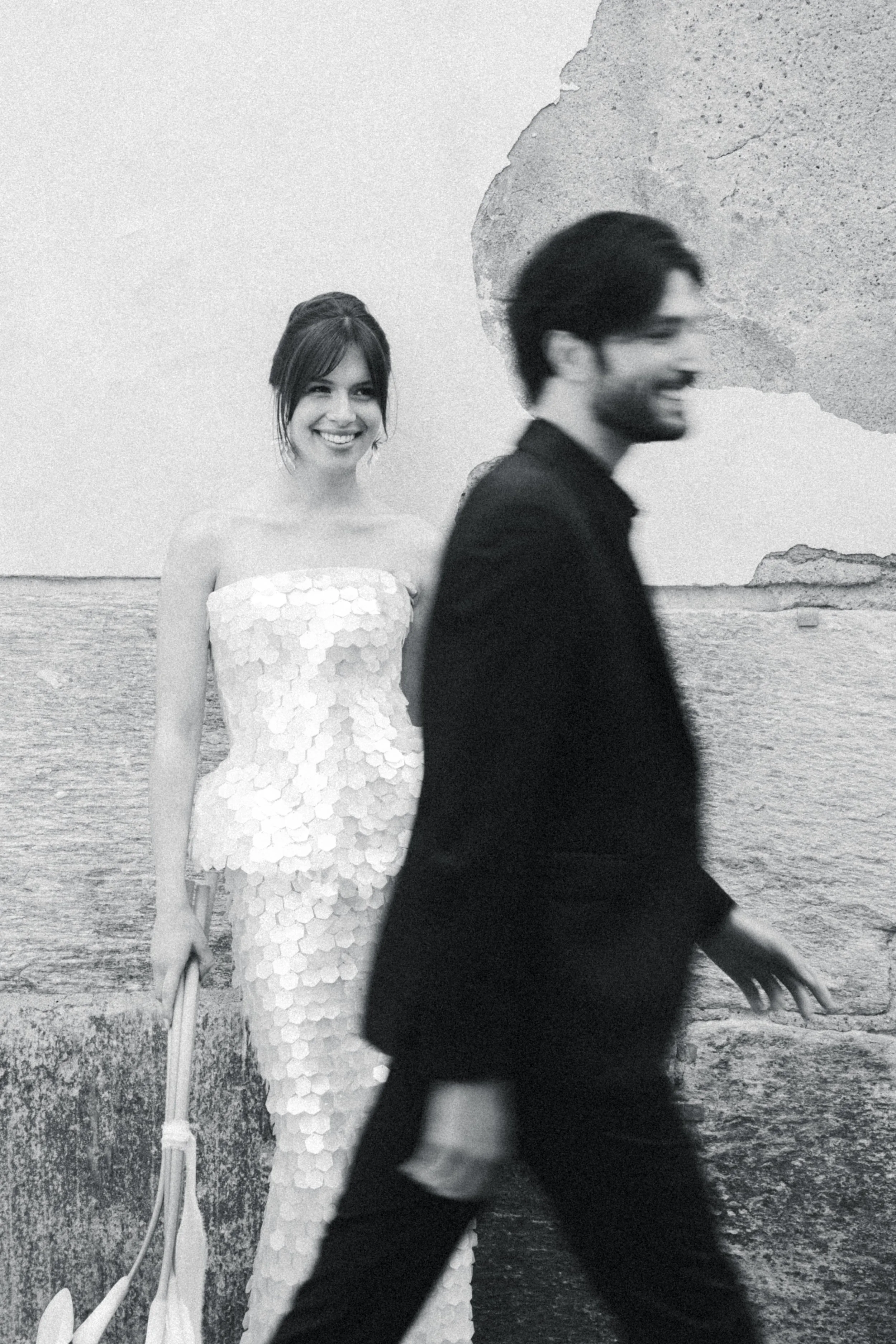 Black and white photo of a woman in a wedding dress smiling, with a man in a suit walking past in front of her by the beach, with rocks in the background.