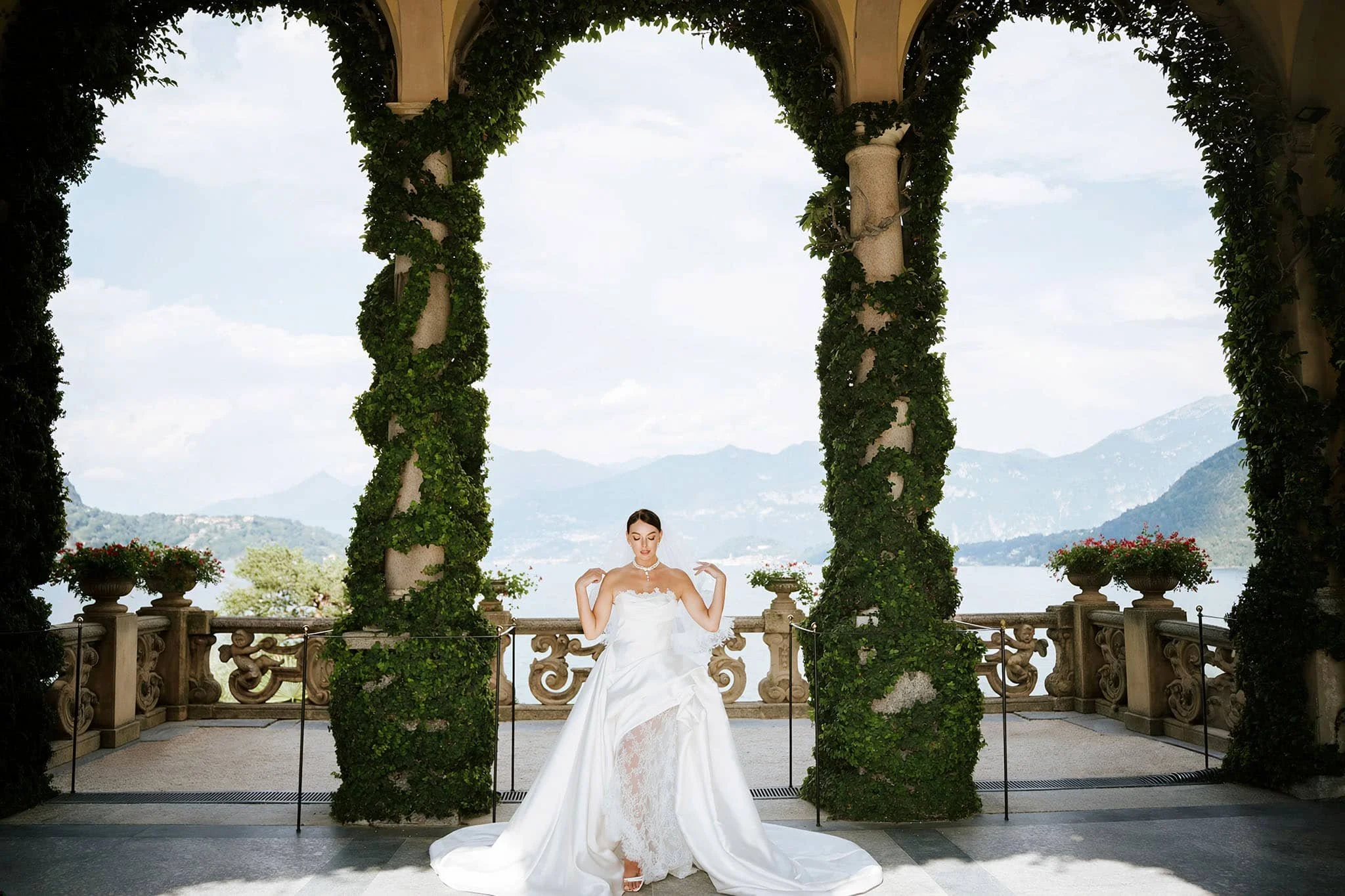Bride sitting on a stone bench under the ivy-covered loggia arches at Villa del Balbianello, Lake Como, with mountain views behind her.