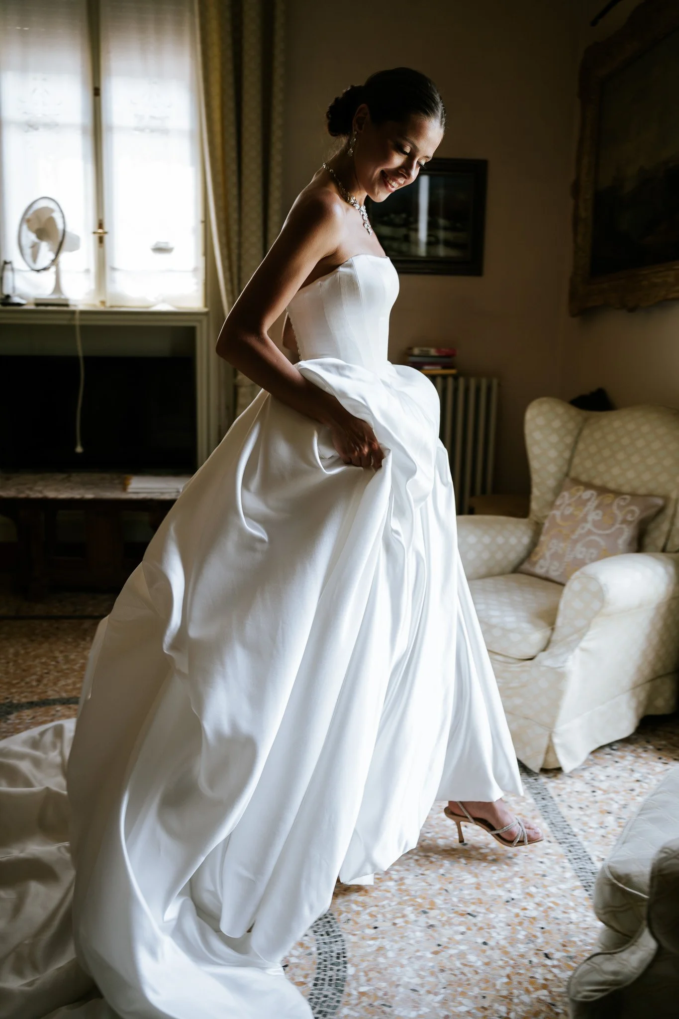 A woman in a wedding dress smiling and adjusting her gown inside a room with a patterned carpet and cushioned furniture.