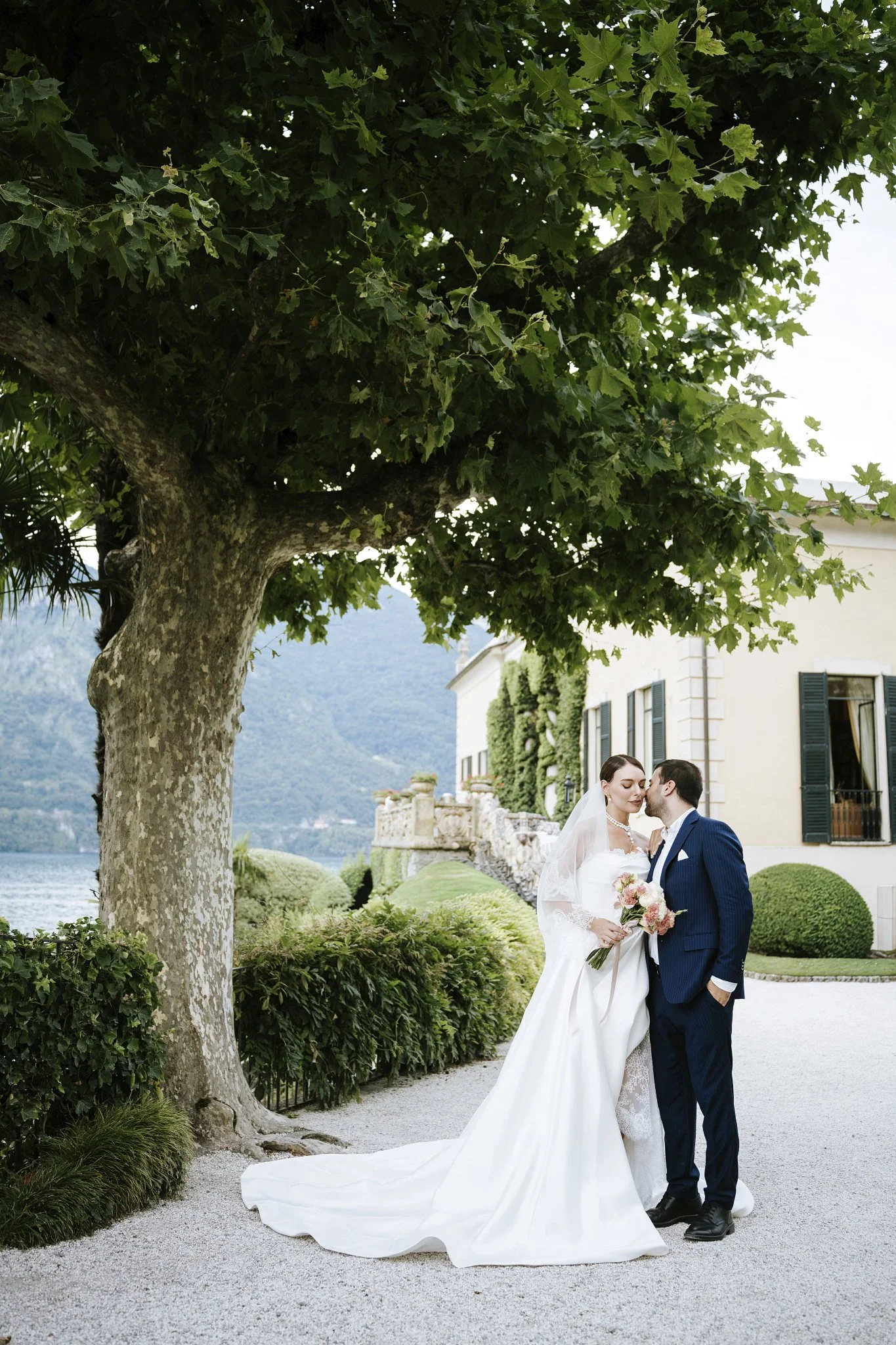Bride and groom under a tree in the gardens of Villa del Balbianello, Lake Como, with the lake and mountains behind them.
