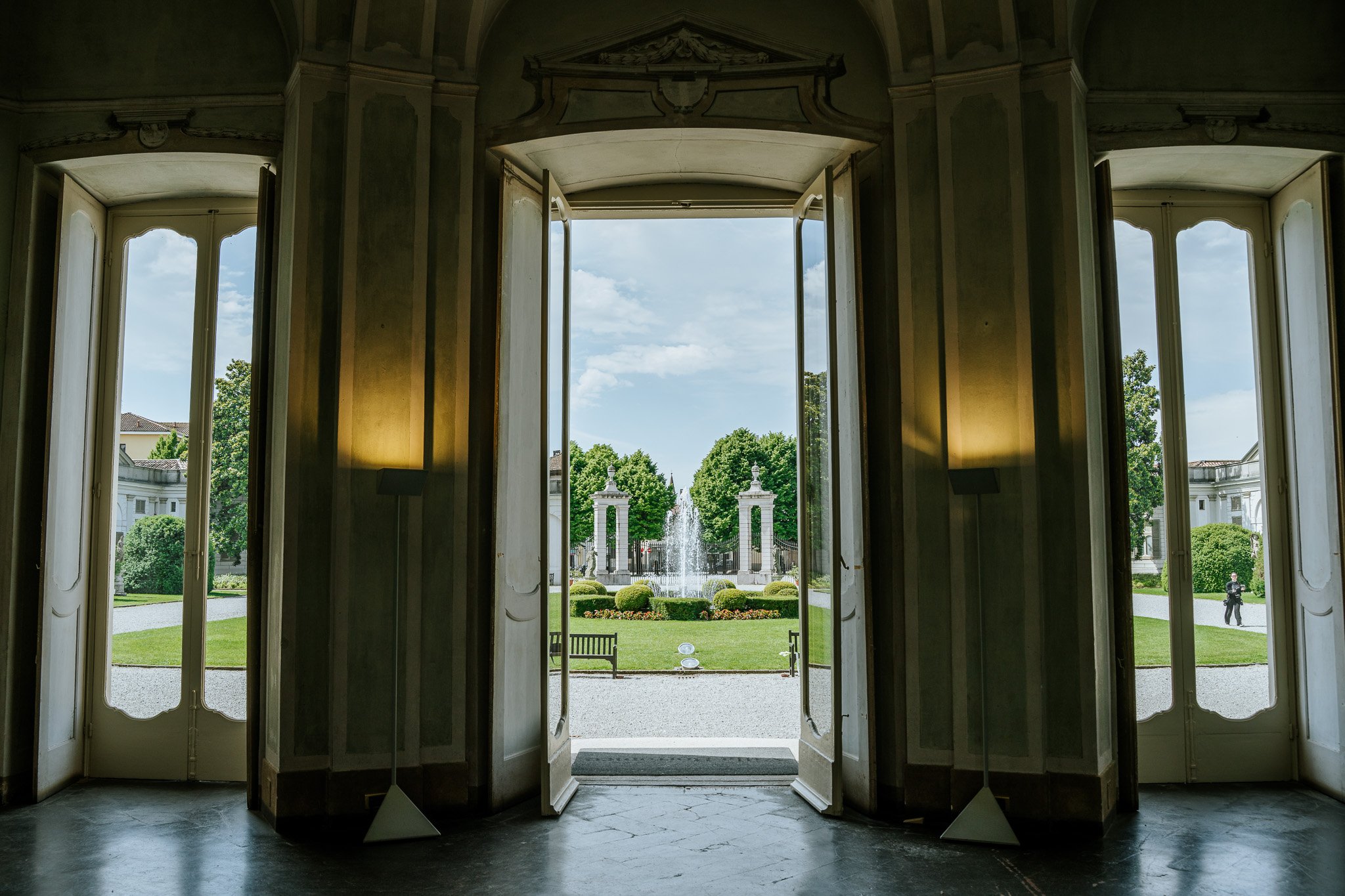 Open double doors leading to a garden with a fountain, green trees, and white classical architecture in the background.