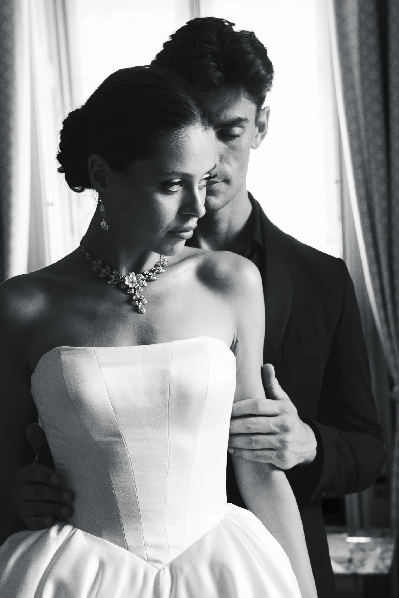 A black and white photo of a woman in a strapless wedding gown and jewelry, with a man in a suit behind her, standing near a window.