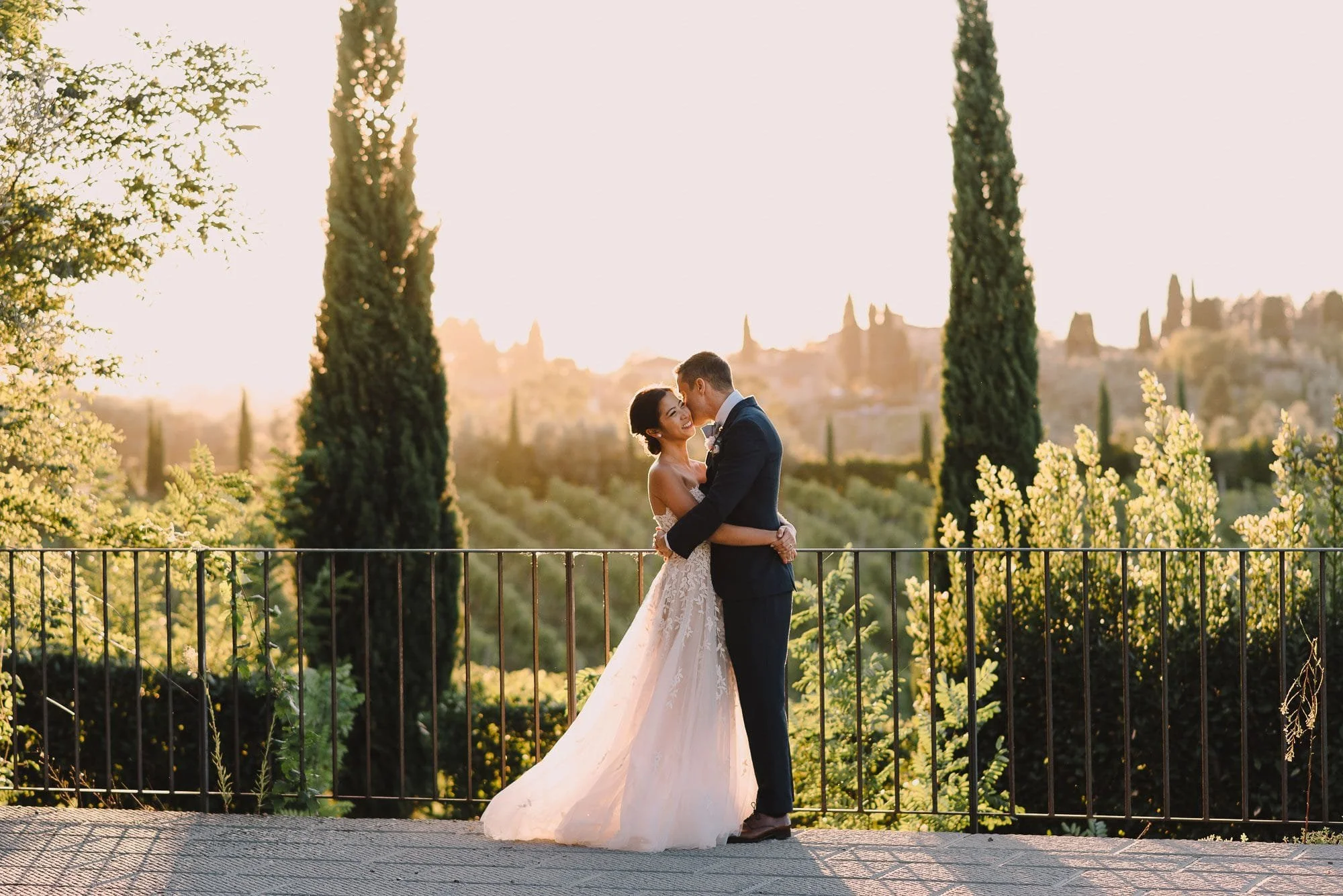 A bride and groom share a kiss during their wedding ceremony outdoors, with tall trees and a scenic view in the background during sunset.