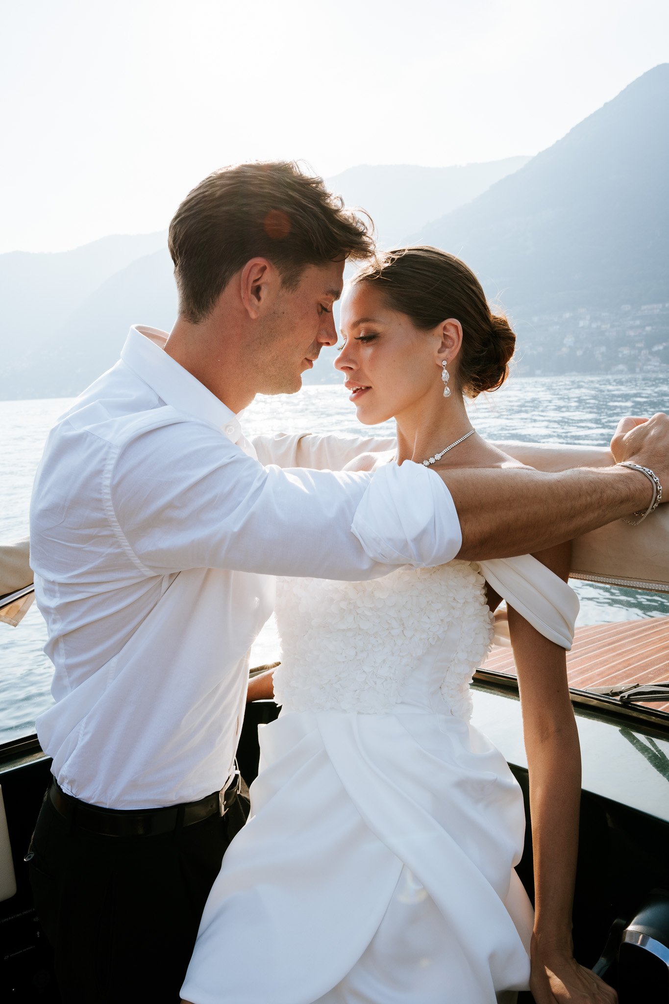 A man and woman dressed in wedding attire embracing on a boat with a water and mountain landscape in the background.