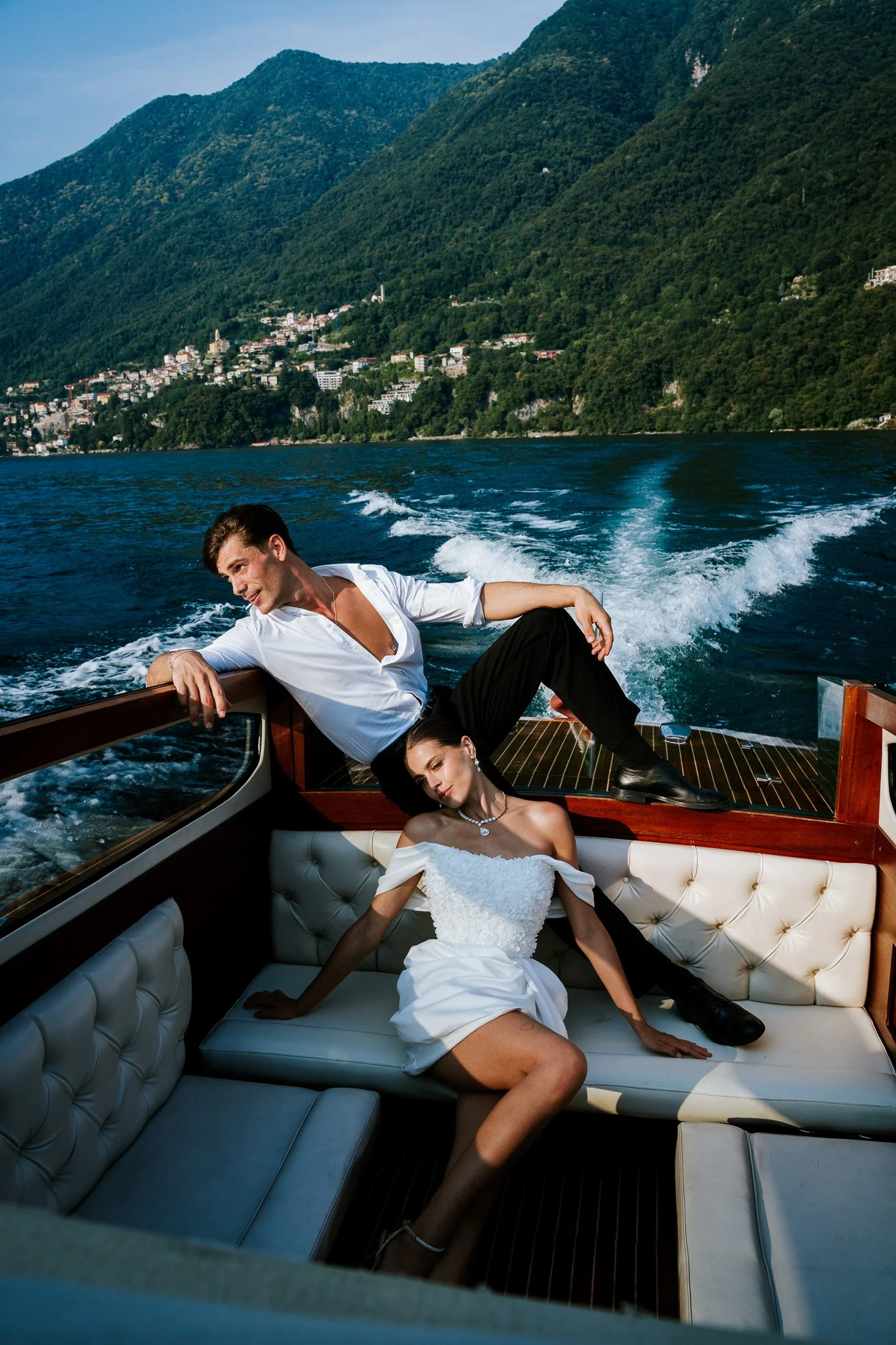 A man in a white shirt and black pants is leaning on the side of a yacht, smiling and looking away. A woman in a white dress and jewelry is lying on the cushioned seating inside the yacht, with her eyes closed. The yacht is on a body of water with mo