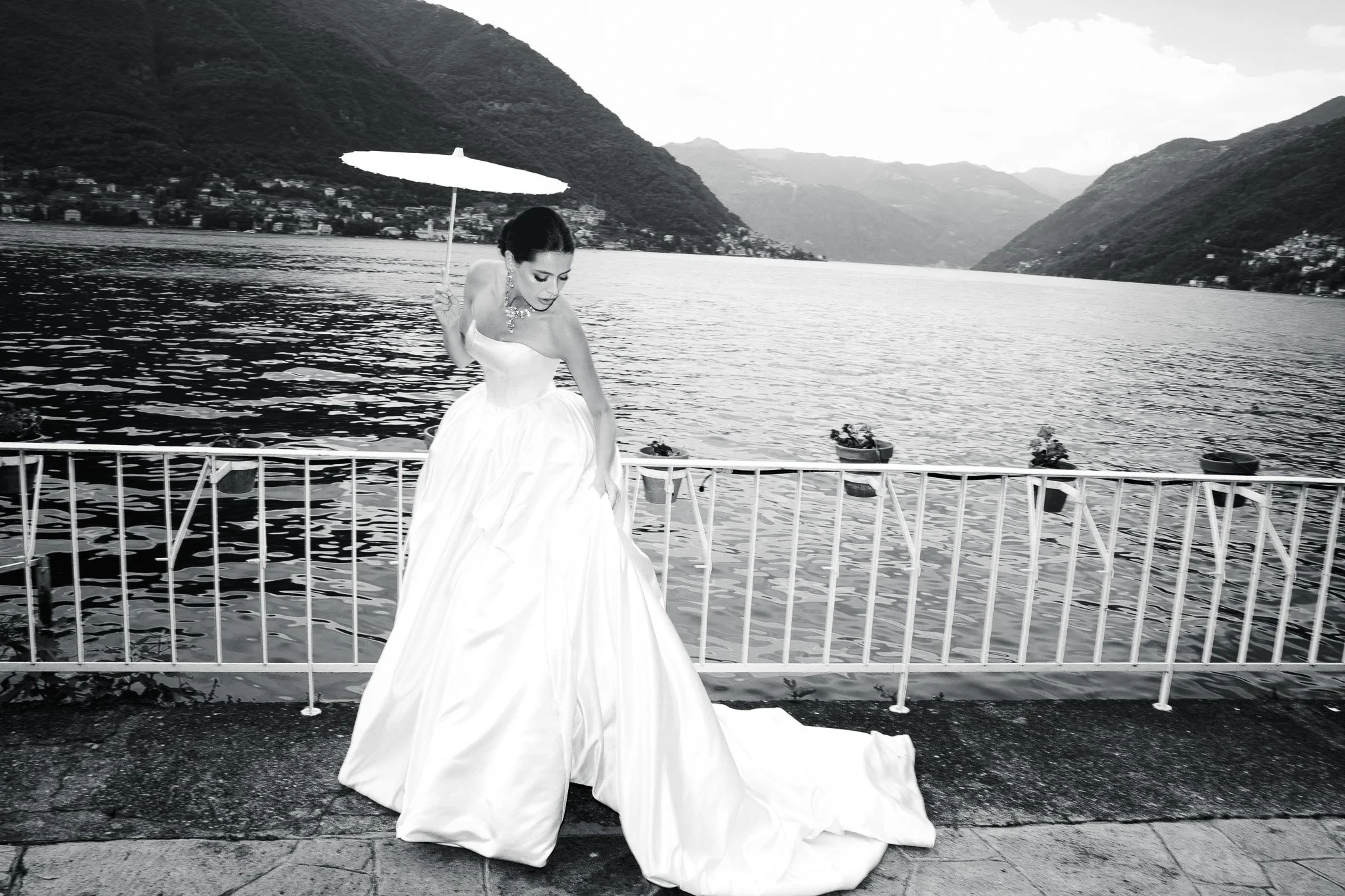 A woman in a wedding dress holding a white parasol by a lake with mountains in the background.
