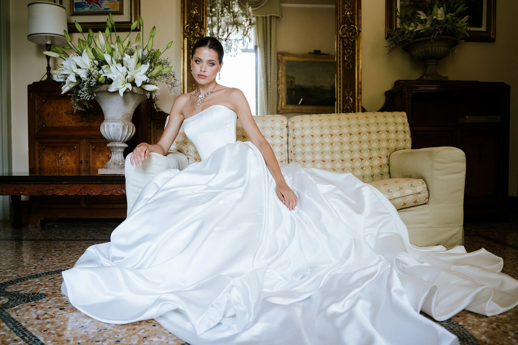 A woman wearing a white wedding dress sitting on a cream-colored couch in an elegant room with large floral arrangements, a mirror, and framed artwork.
