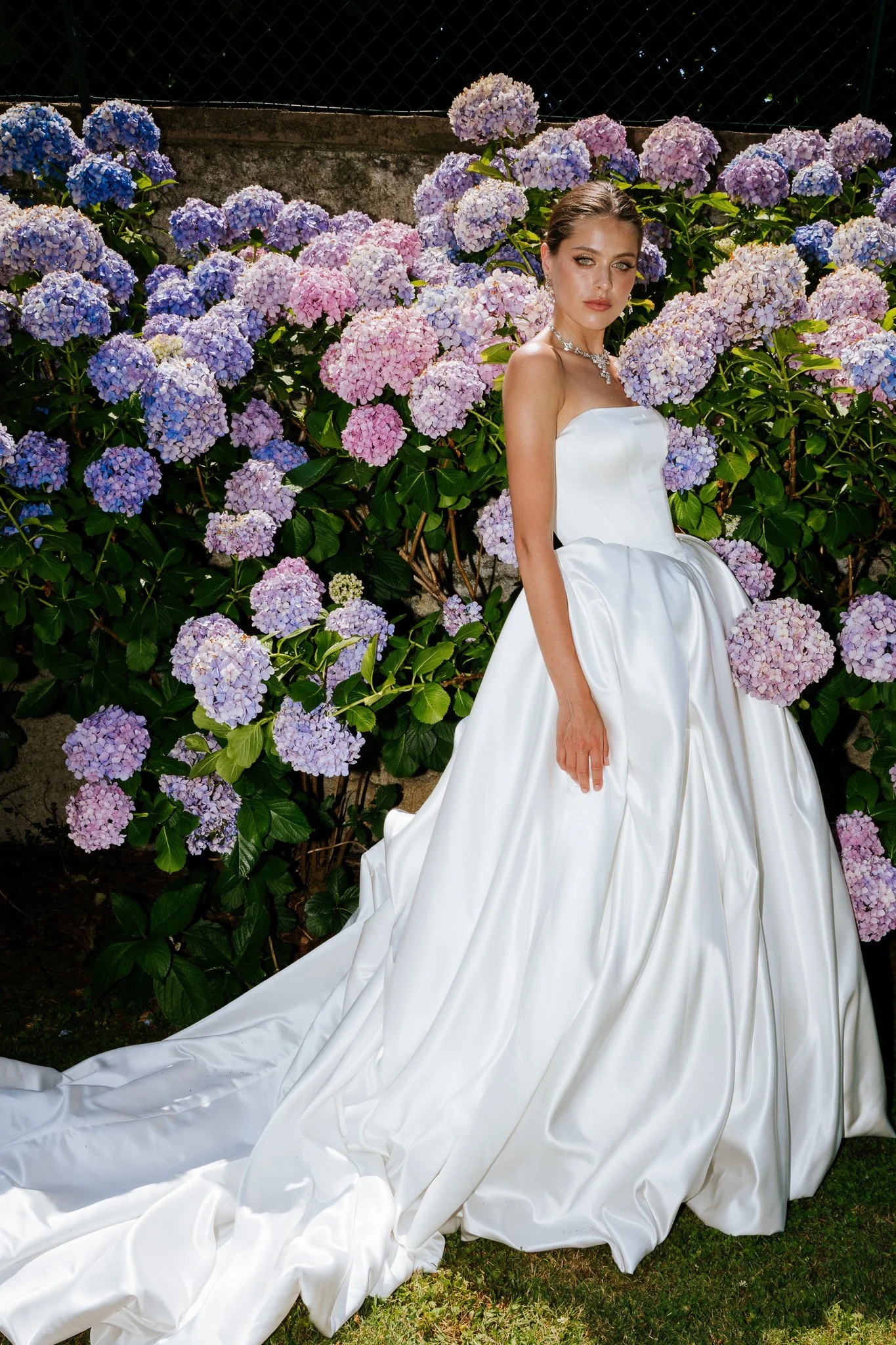 A woman in a white strapless wedding gown with a long train standing in front of purple and pink hydrangea flowers.