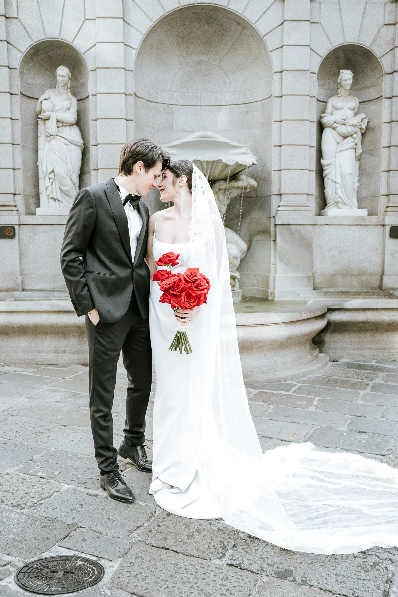 A newlywed couple standing close, touching foreheads, outdoors near a stone fountain with sculptures, one holding a bouquet of red roses, both dressed in wedding attire.