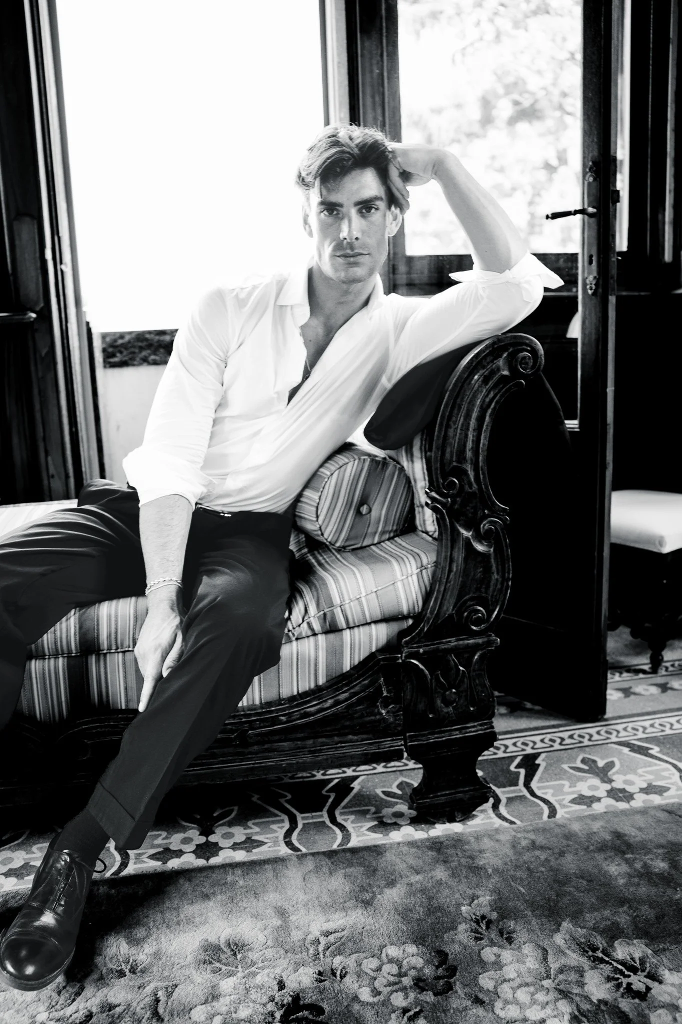 A young man with styled hair, wearing a white shirt and dark pants, sitting on an ornate wooden sofa with striped cushions near a large window, with a patterned rug on the floor in a room with natural light.