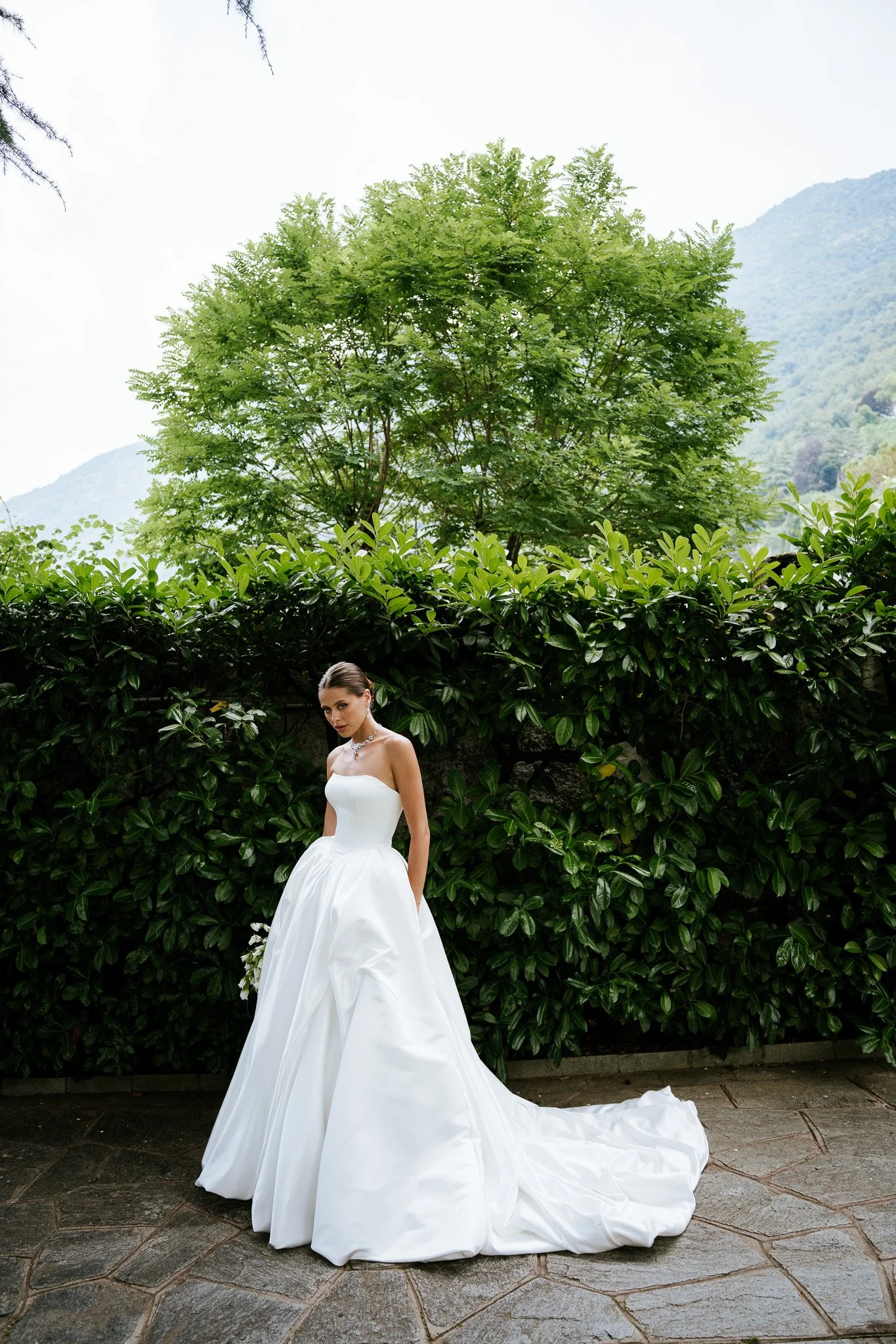 A woman in a white wedding gown standing outdoors in front of dense green foliage, with mountains in the background.
