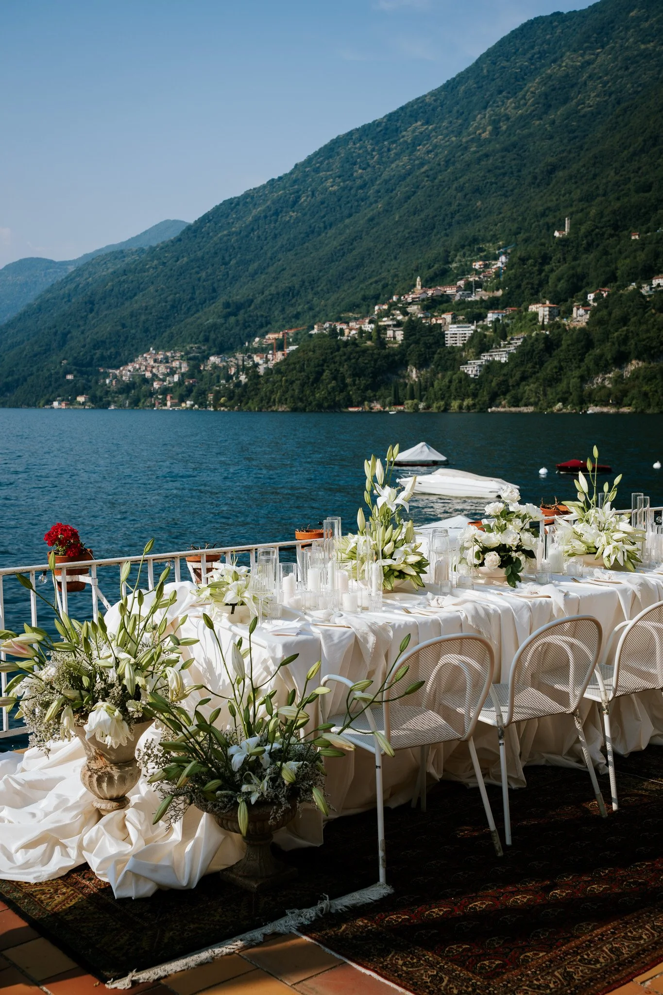 A long, elegant banquet table set for an outdoor event by a lake with mountains in the background. The table is decorated with white linens, glassware, and white floral centerpieces. There are chairs around the table, and the scene looks tranquil and