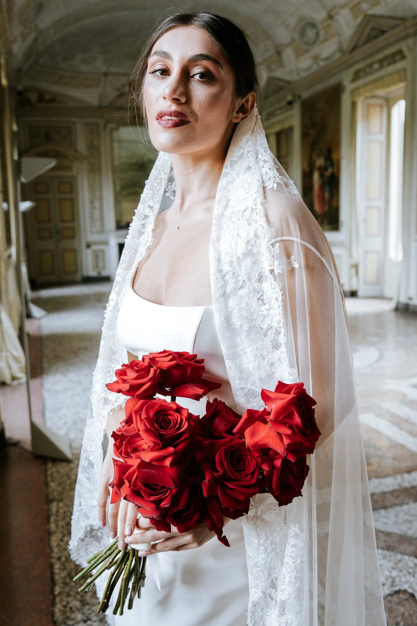A woman wearing a wedding dress and lace veil, holding a bouquet of red roses, standing inside a historic, ornately decorated room with tall windows and intricate ceiling details.