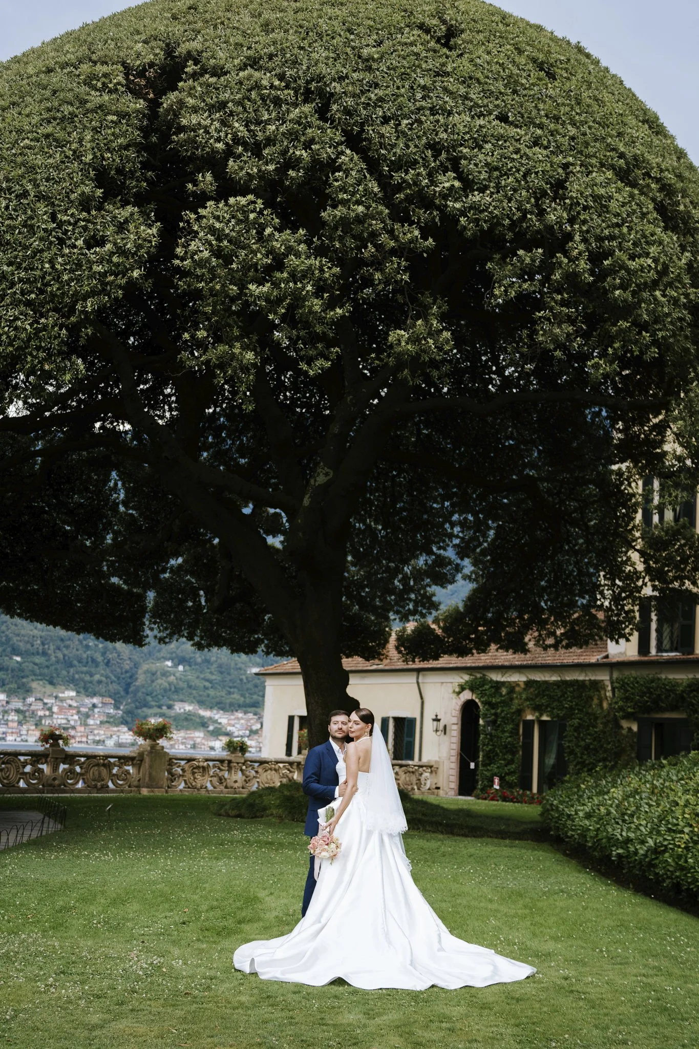 Bride and groom under a tree at Villa del Balbianello, Lake Como, the bride holding a bouquet of pink flowers.