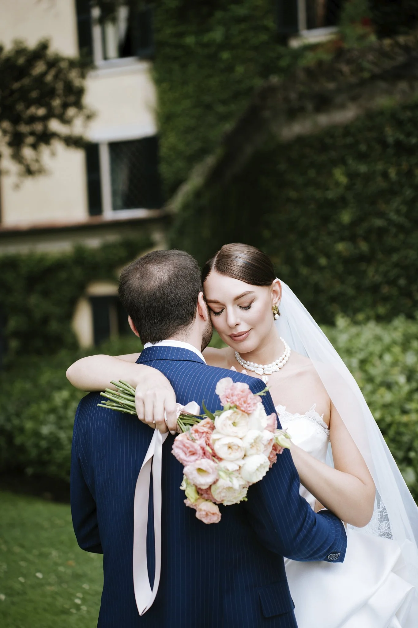 Bride and groom embracing at Villa del Balbianello, Lake Como, the bride holding a bouquet of pink and white roses.