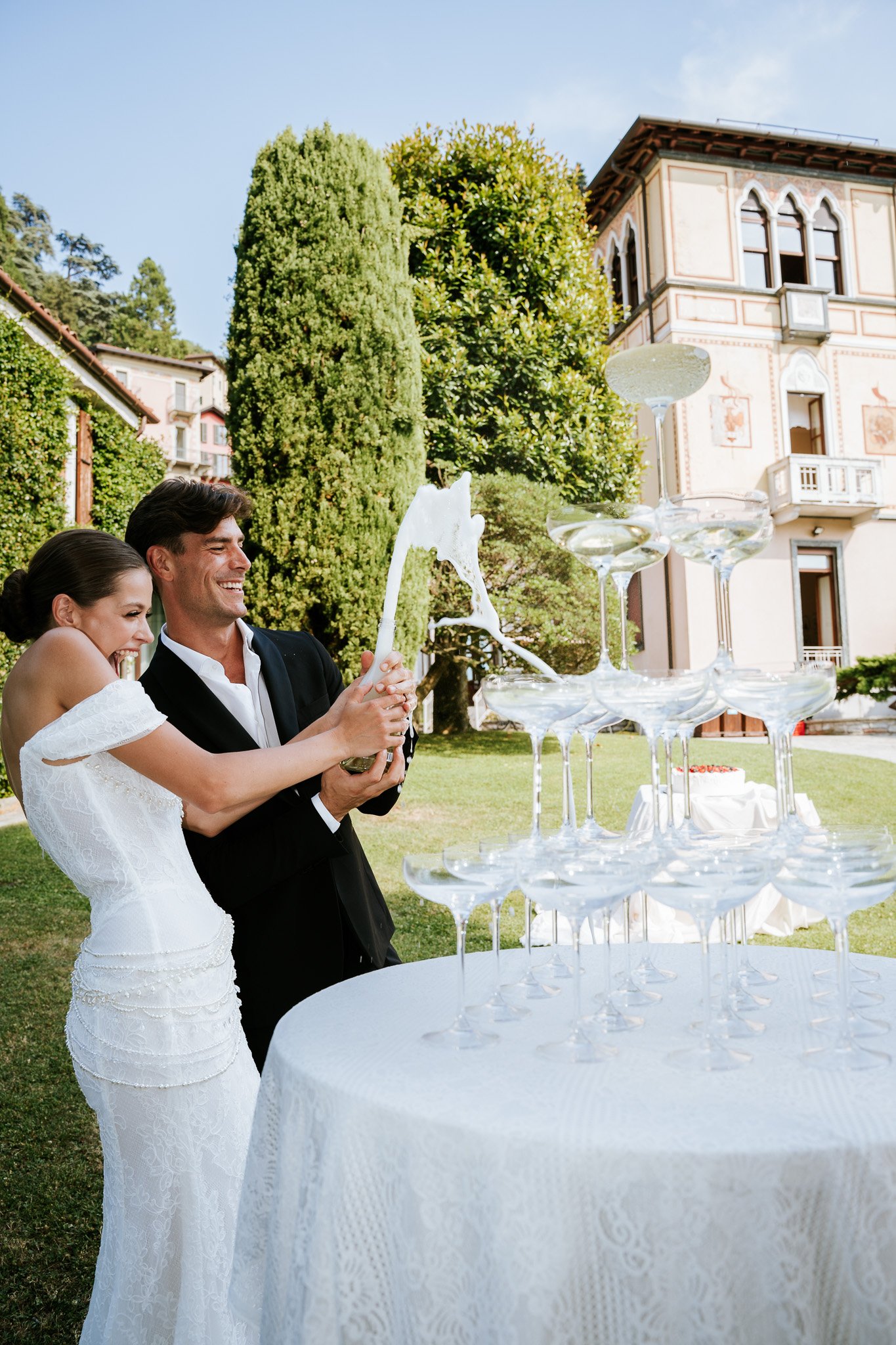 A bride and groom at a wedding celebration outdoors, standing near a dessert display with champagne glasses and a tower of wine glasses containing water, smiling and holding a champagne flute together.