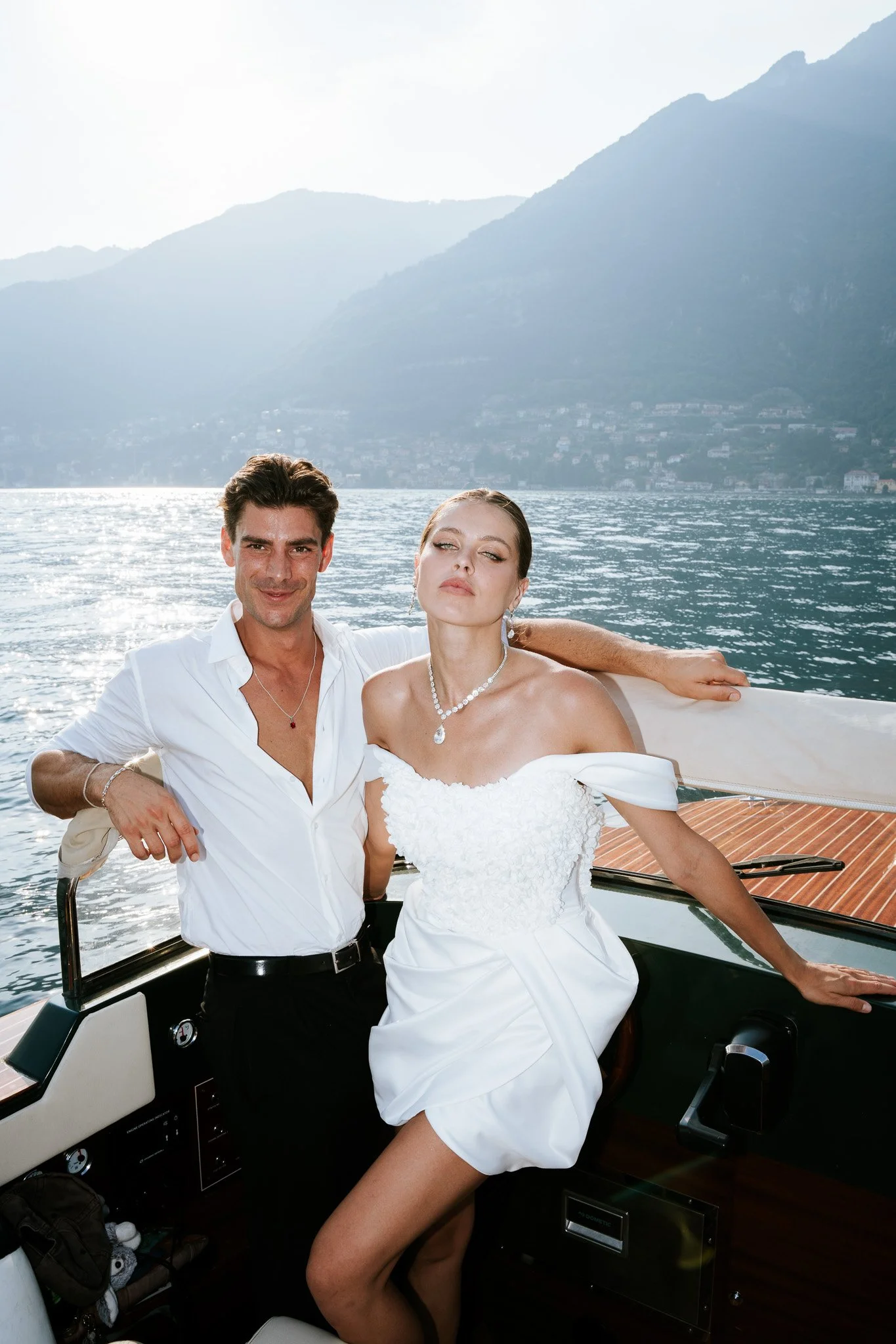 A man and woman on a boat with a lake and mountains in the background. The man is smiling slightly and wearing a white shirt, and the woman is wearing a white off-shoulder dress and jewelry, relaxing with her arm around the man.