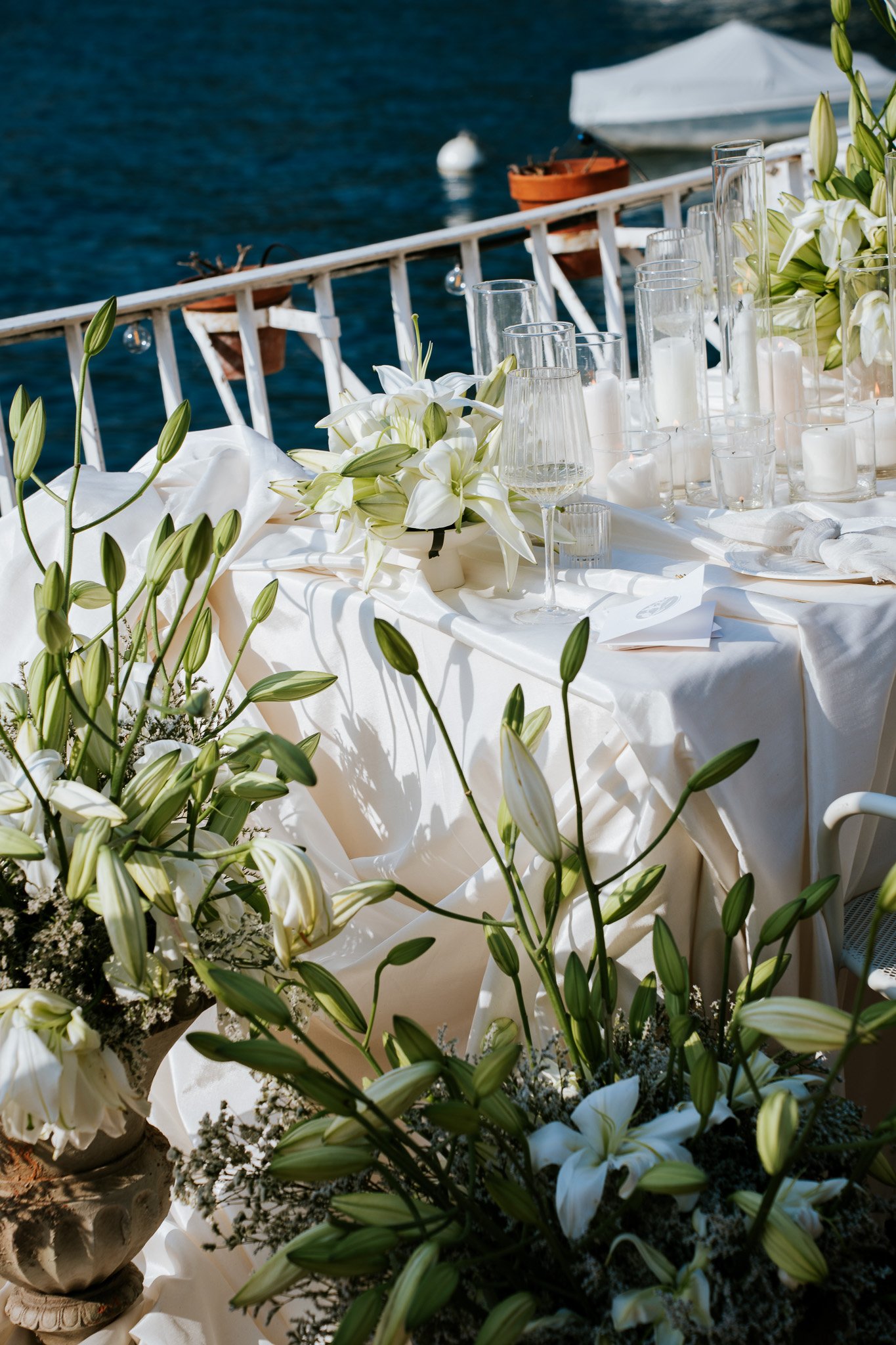 A beautifully decorated table with white lilies and candles, set near a body of water with a boat in the background.