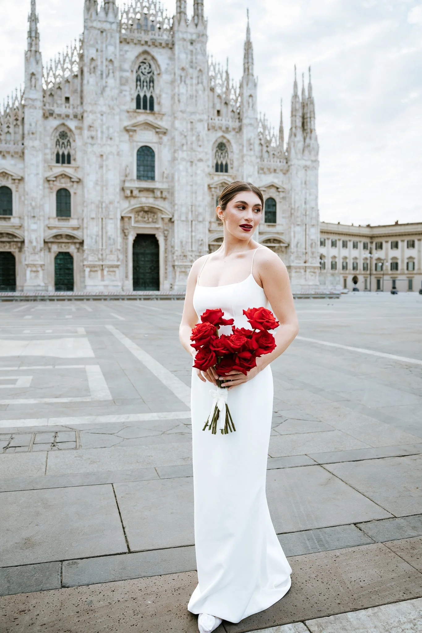 A woman in a white wedding dress holding a bouquet of red roses, standing in front of a historic cathedral with pointed spires and ornate stonework.