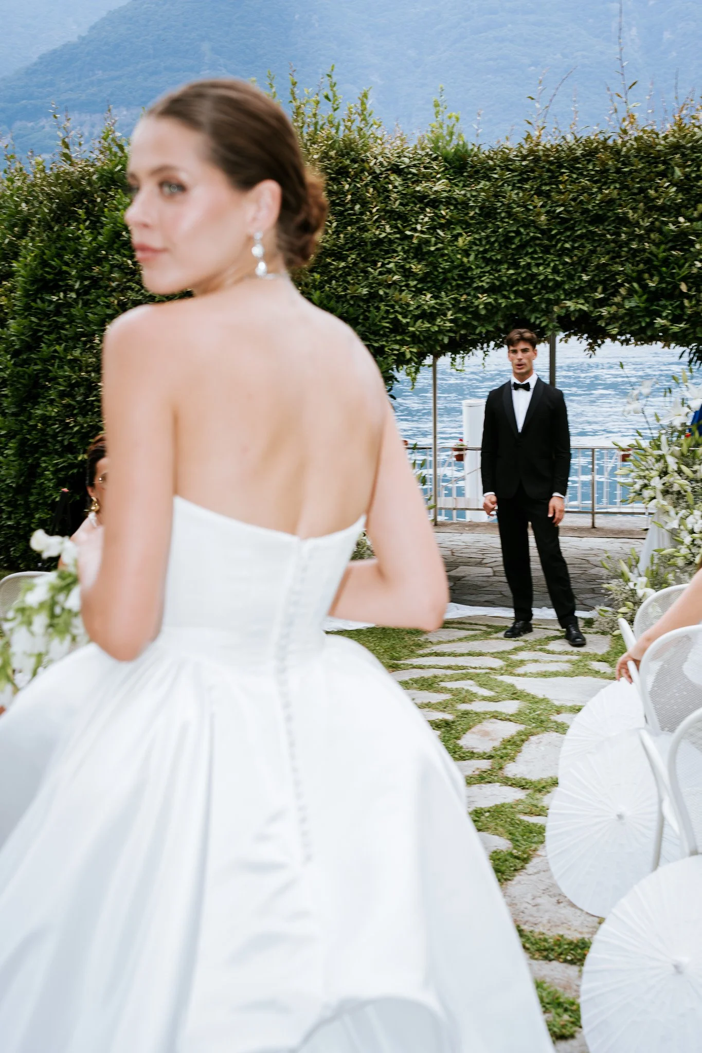 A woman in a white wedding dress with brown hair styled in an updo, wearing earrings, facing left, in the foreground. In the background, a man in a black tuxedo with a bow tie stands near water, with greenery and mountains behind him, at an outdoor w