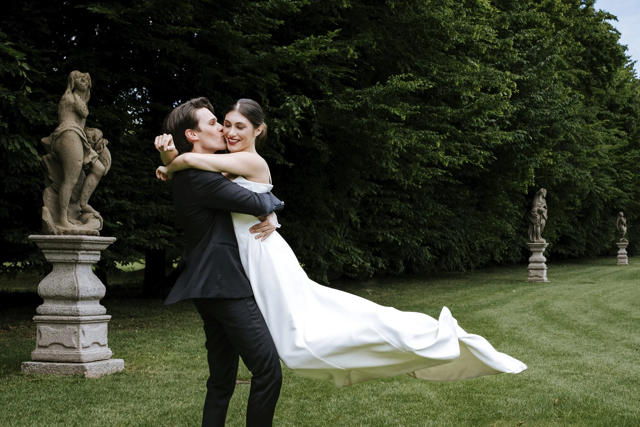 Bride and groom sharing a kiss in a historic Lake Como villa garden surrounded by statues and lush greenery.